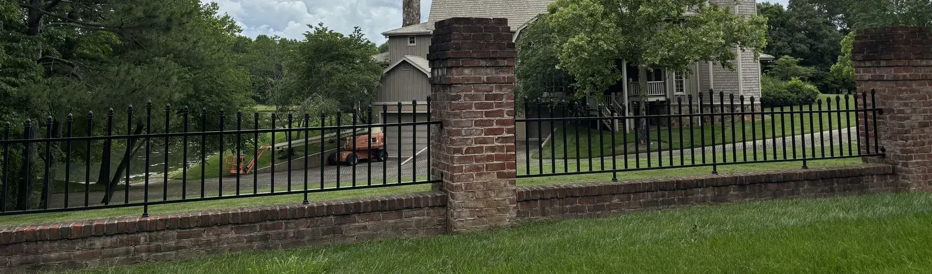 Brick pillar and black metal fence surround a grassy area with trees and buildings in the background.