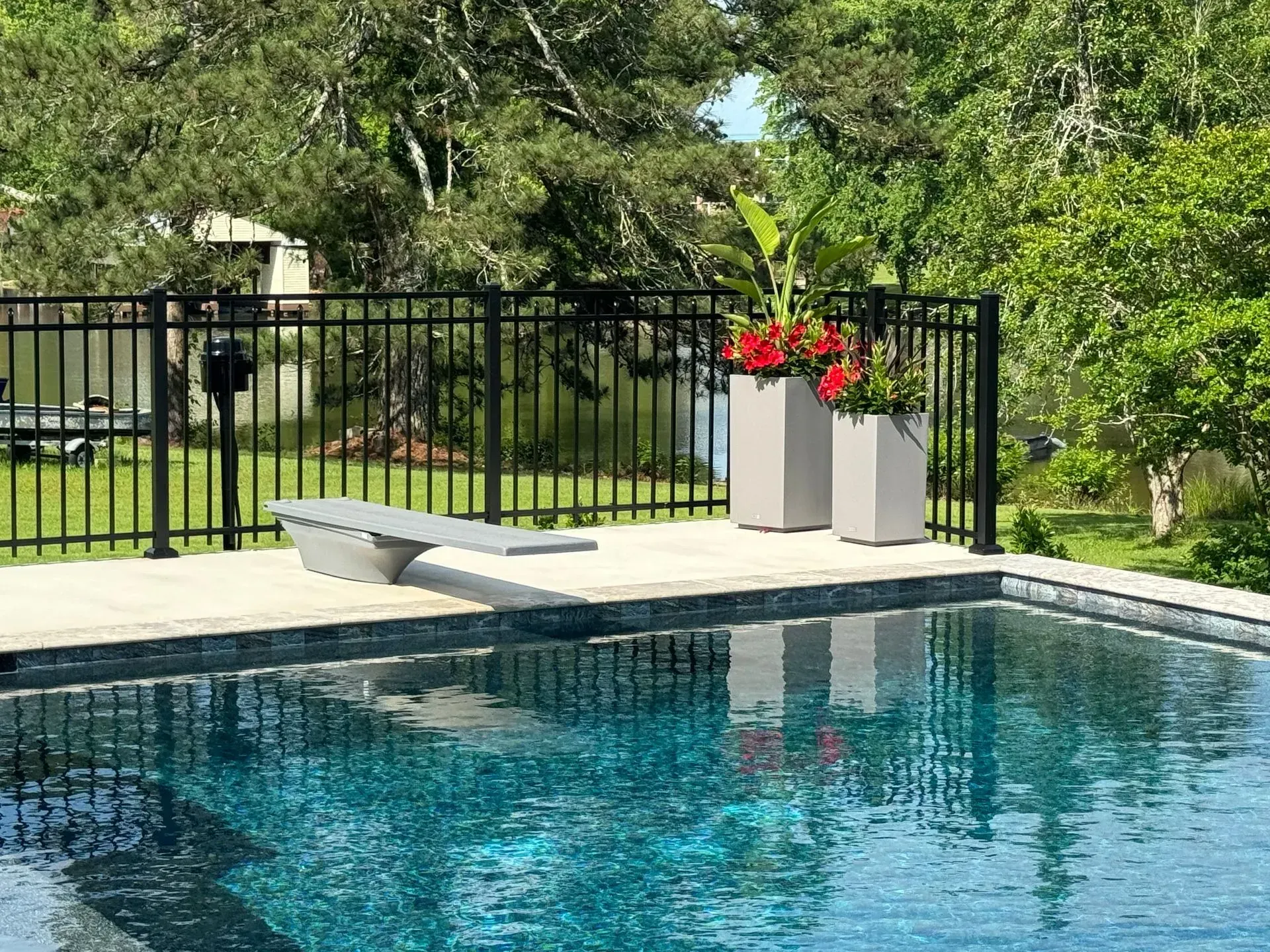 Swimming pool with a diving board, black fence, and two planters with red flowers.