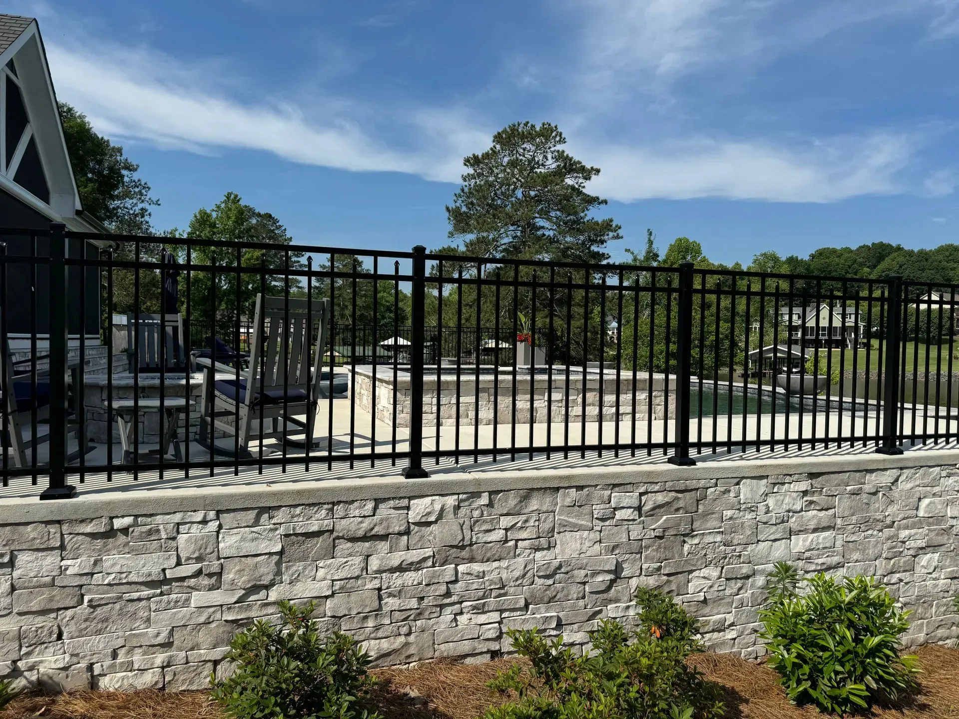 Black metal fence atop a stone wall, surrounding an outdoor area with trees and sky.