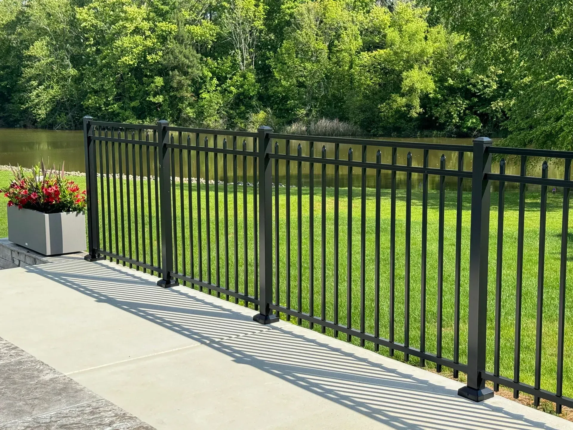 Black metal fence on concrete patio, green grass, trees, and water in background.