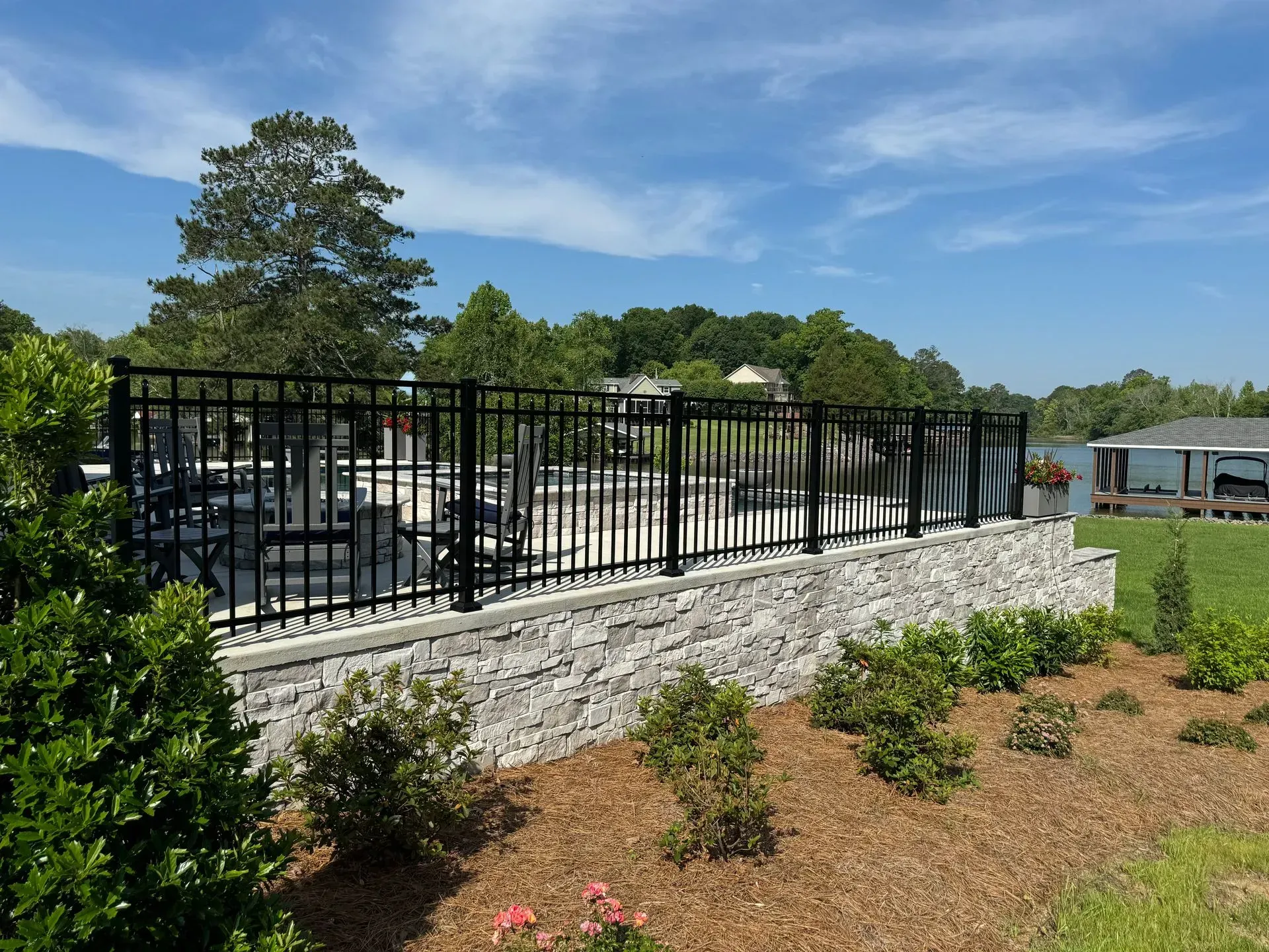 Black metal fence atop a stone wall surrounding a pool area. Green landscaping, blue sky, and a lake in the background.