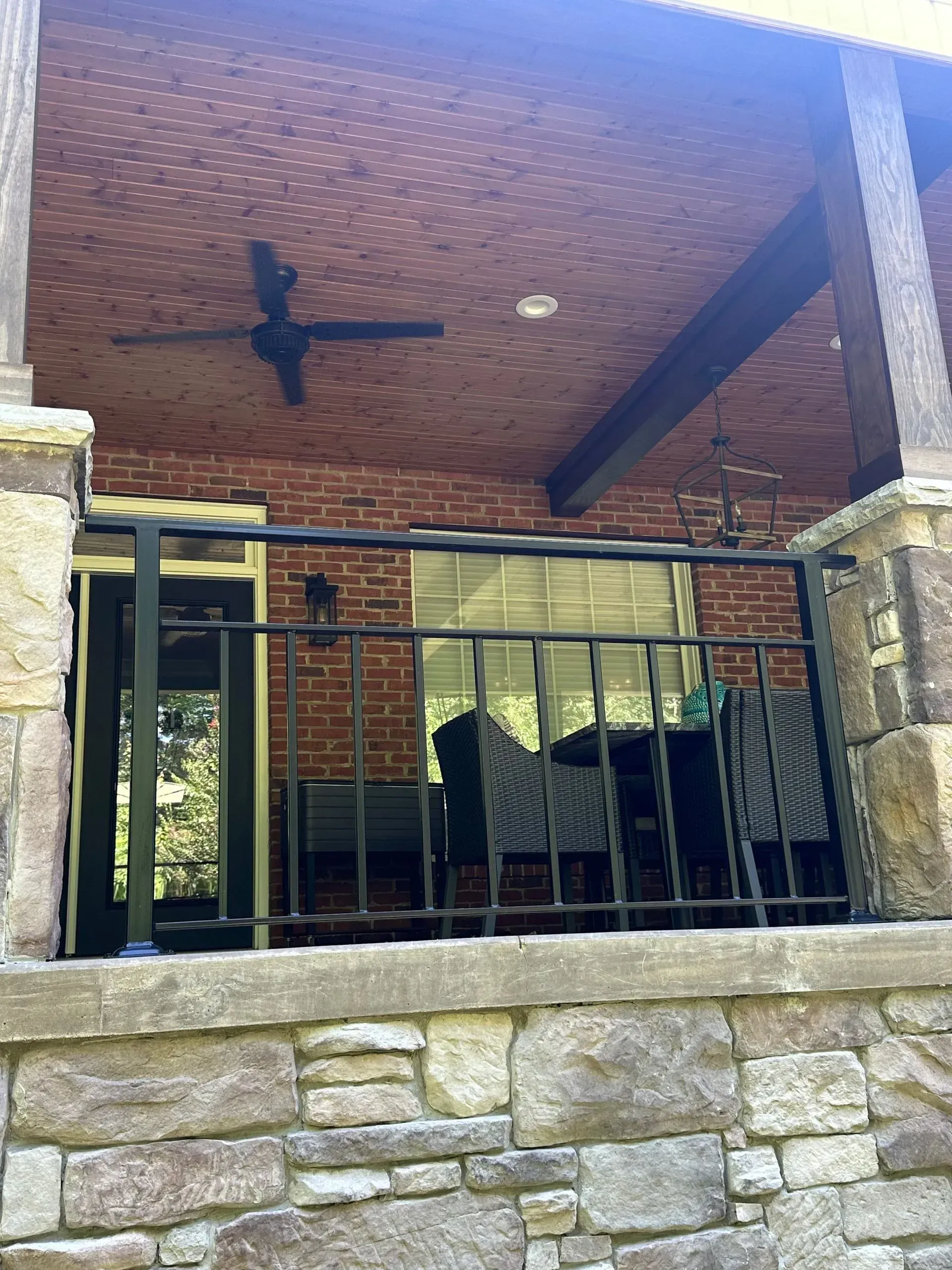 Outdoor patio with brick ceiling, black railing, and stone pillars. Furnished with chairs.