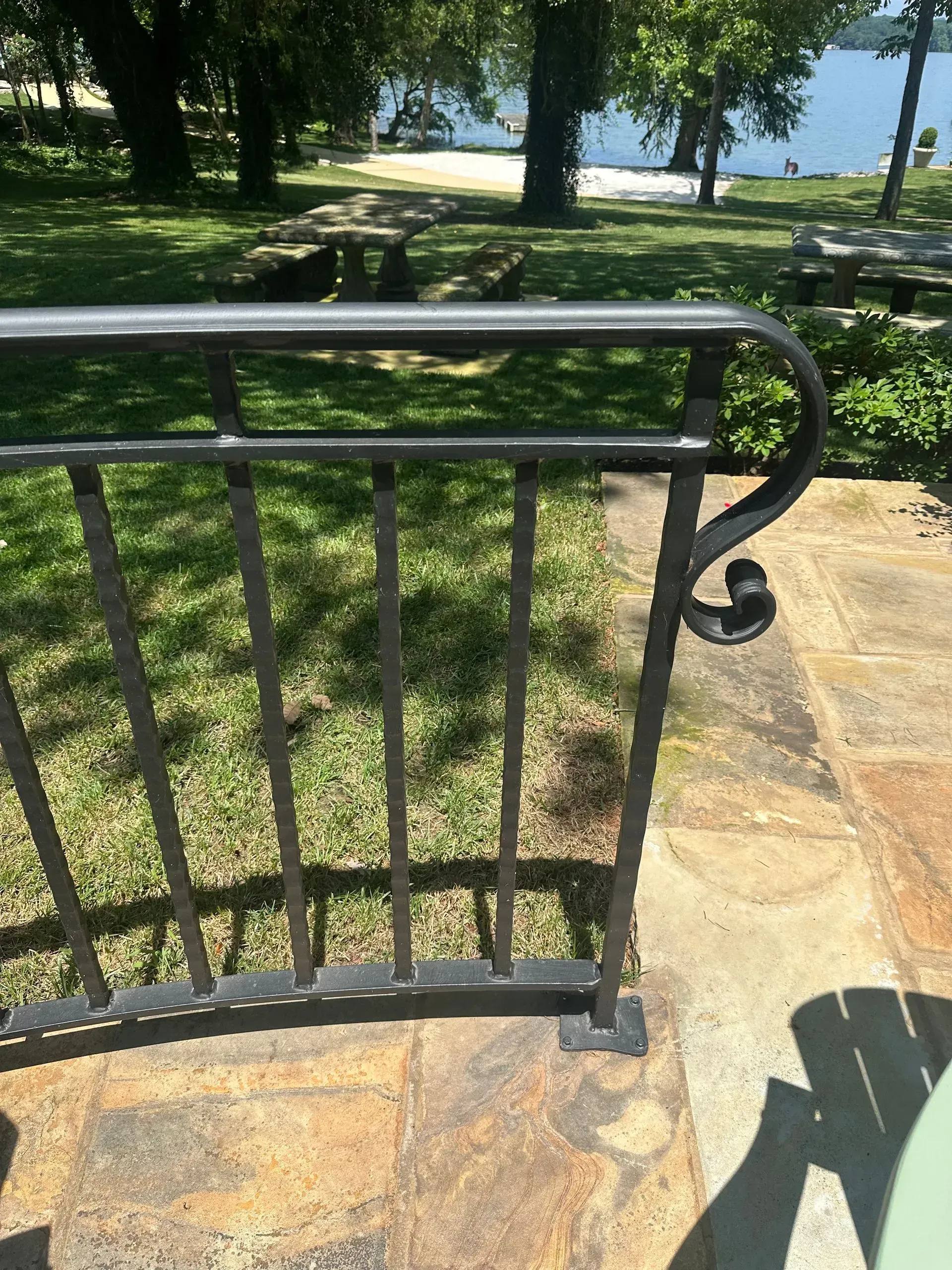 Metal railing on stone patio, overlooking a grassy area and lake with trees.