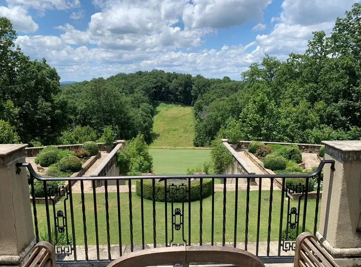 View from a balcony overlooking a green lawn and wooded hillside under a cloudy sky.