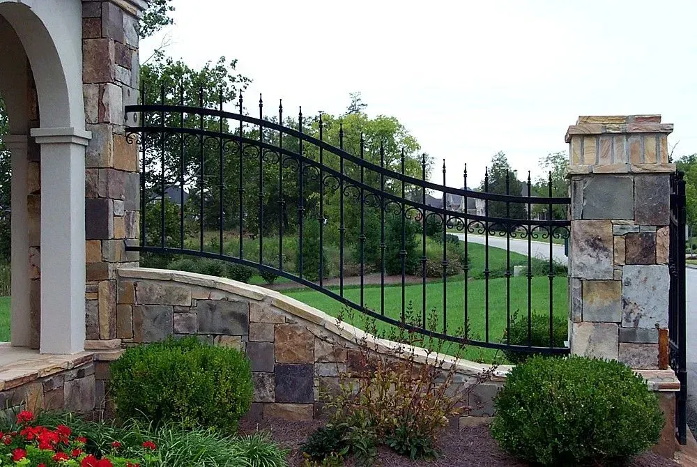 Black wrought iron gate set in a stone wall, curving over a green lawn and road.