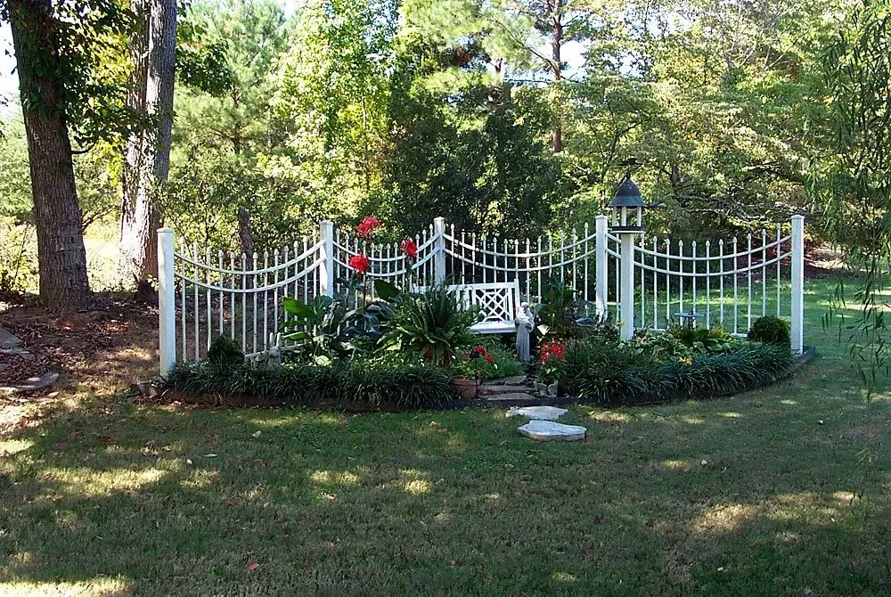 White fenced-in garden with a bench, flowers, and greenery. A lantern sits atop a post. Trees are in the background.