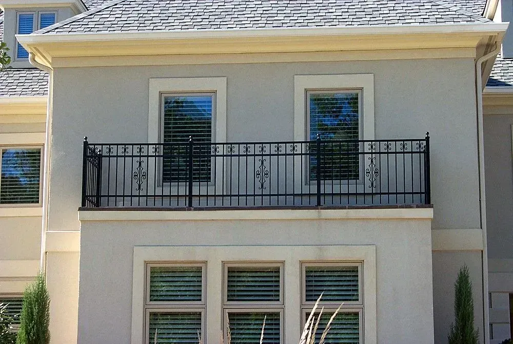 Gray stucco house with black wrought-iron balcony. Two windows with closed blinds on top level, three below.