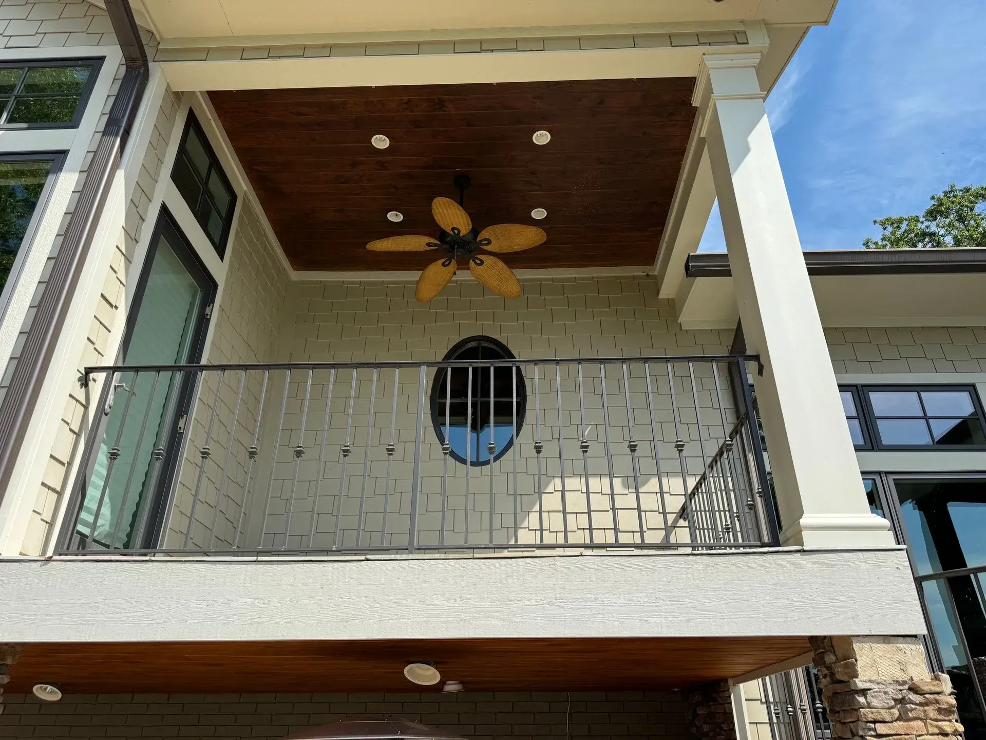 Balcony with metal railing, oval window, ceiling fan, and painted brick exterior under a blue sky.