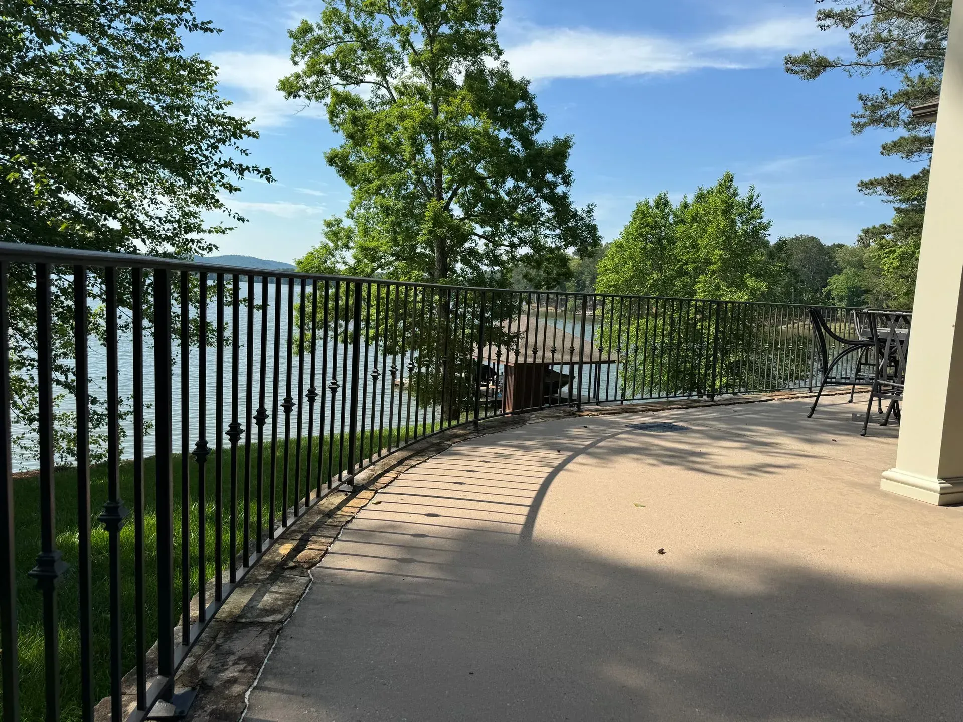 Concrete patio with black metal railing overlooking a body of water with trees, under a blue sky.