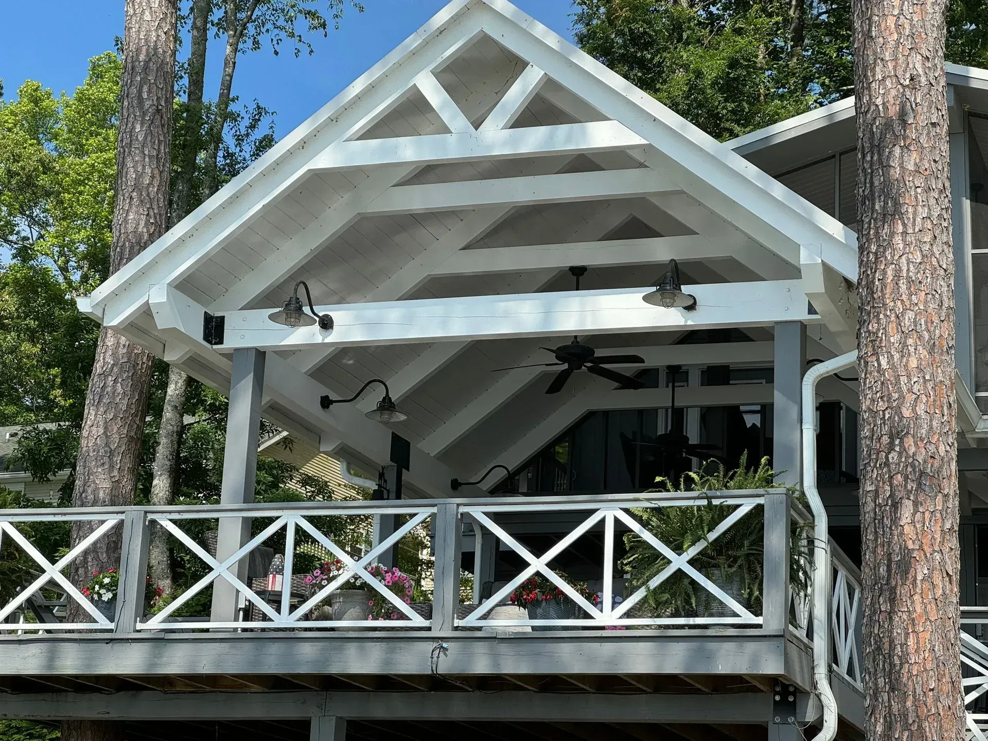 White deck with cross-pattern railing, overhead beams, and gable roof. Trees surround the structure, and it is daytime.