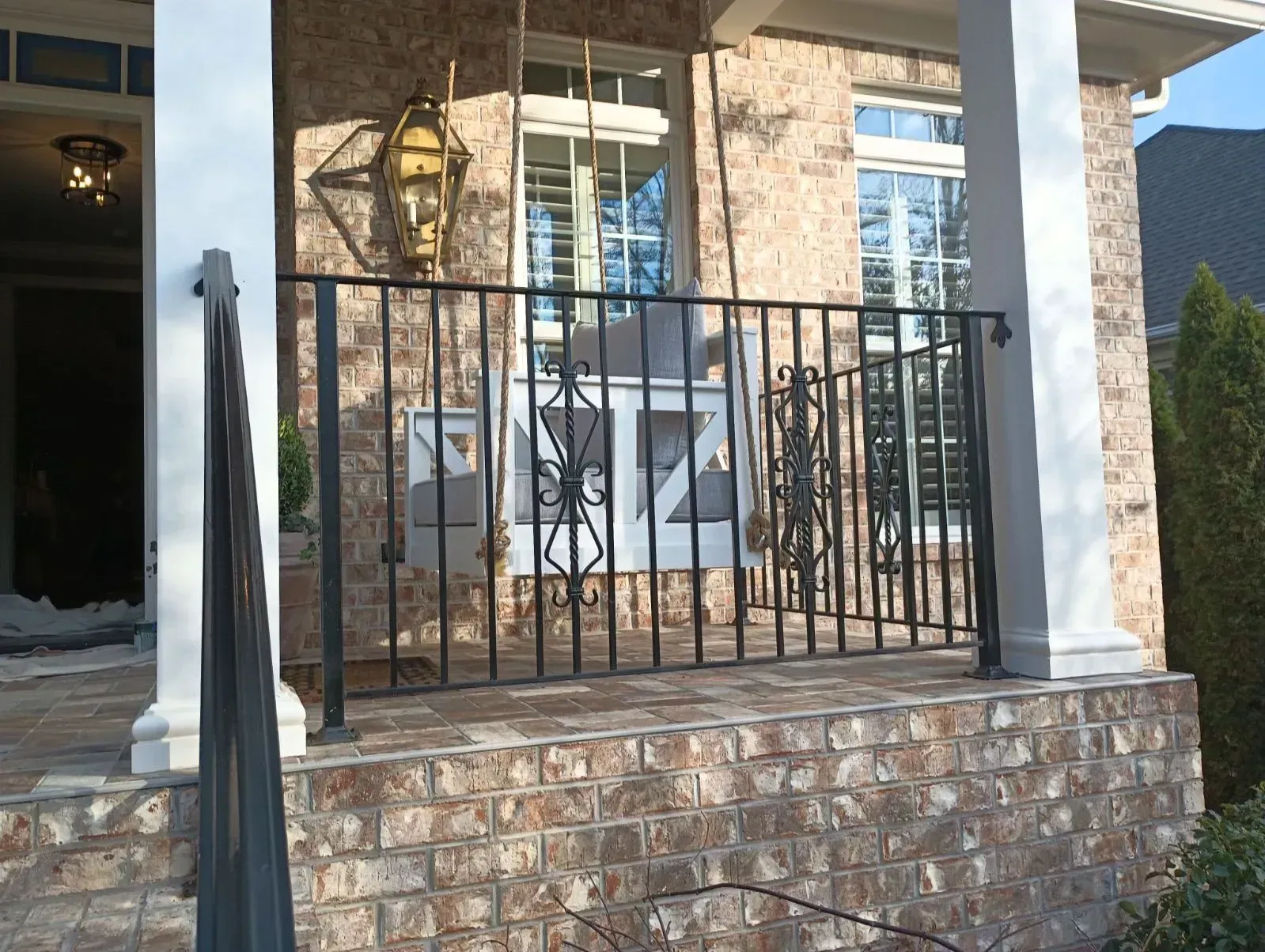 Black wrought-iron railing on brick porch, in front of a house with white columns and windows.