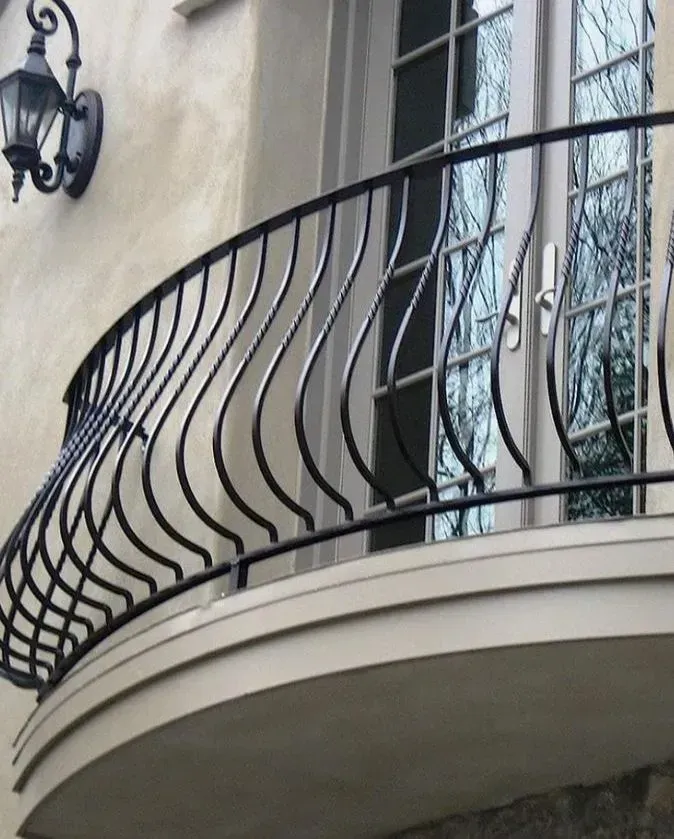 Curved black wrought iron balcony railing on a light-colored building with a door and a sconce.