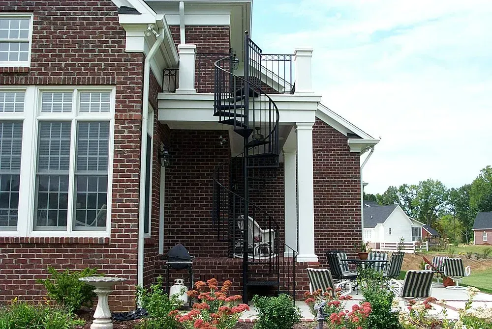 Brick house with black spiral staircase leading to a second-floor balcony. Patio with furniture and garden.
