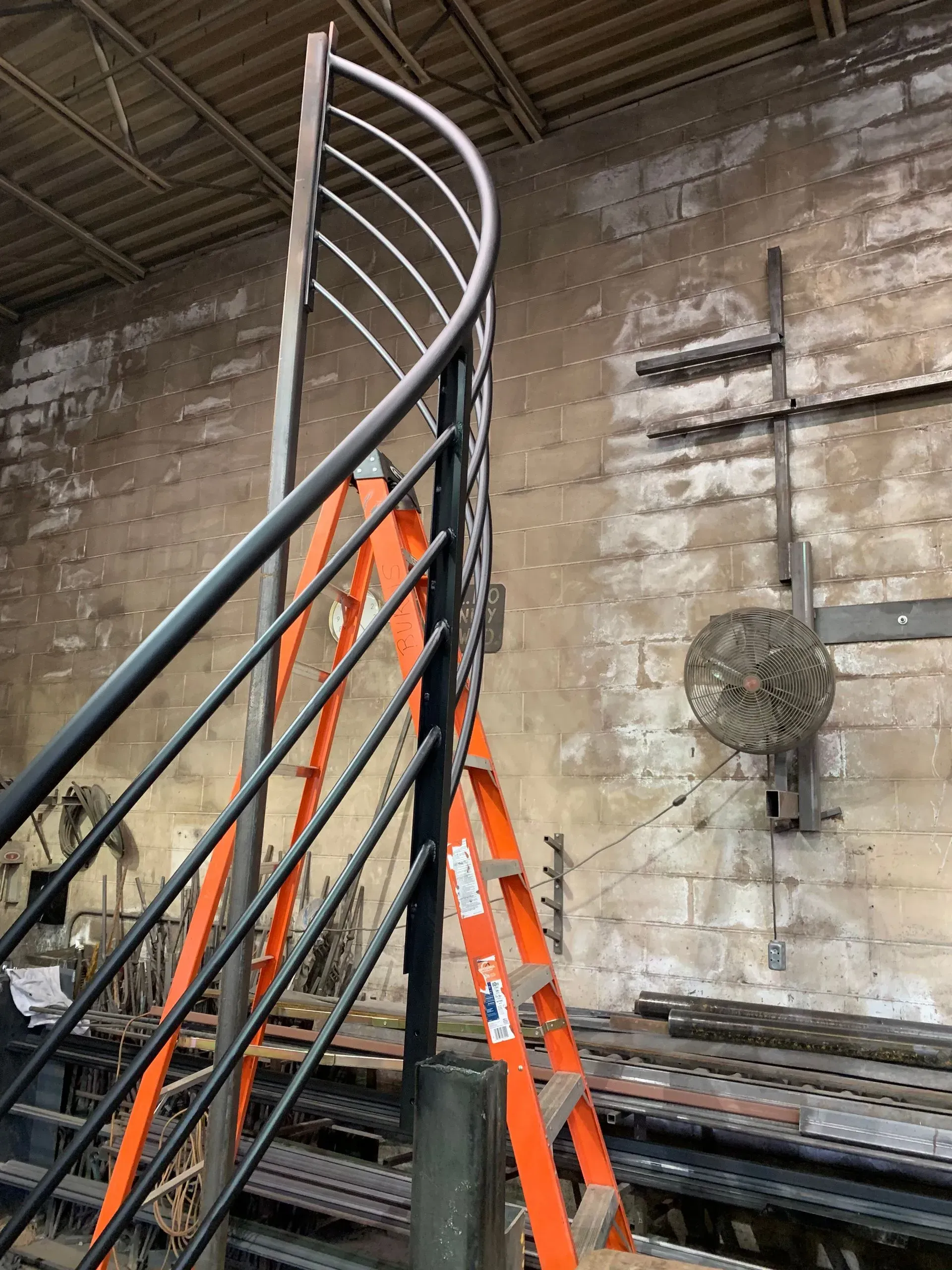 Orange ladder propped against a curved black and silver metal stair railing in an industrial setting.