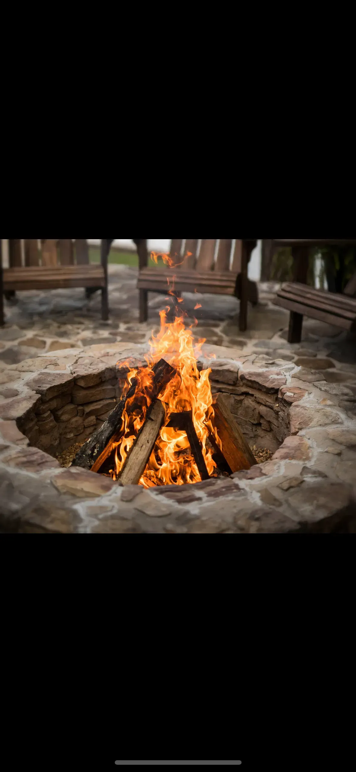 A fire pit with a burning fire, surrounded by a stone border. Wooden chairs are in the background.