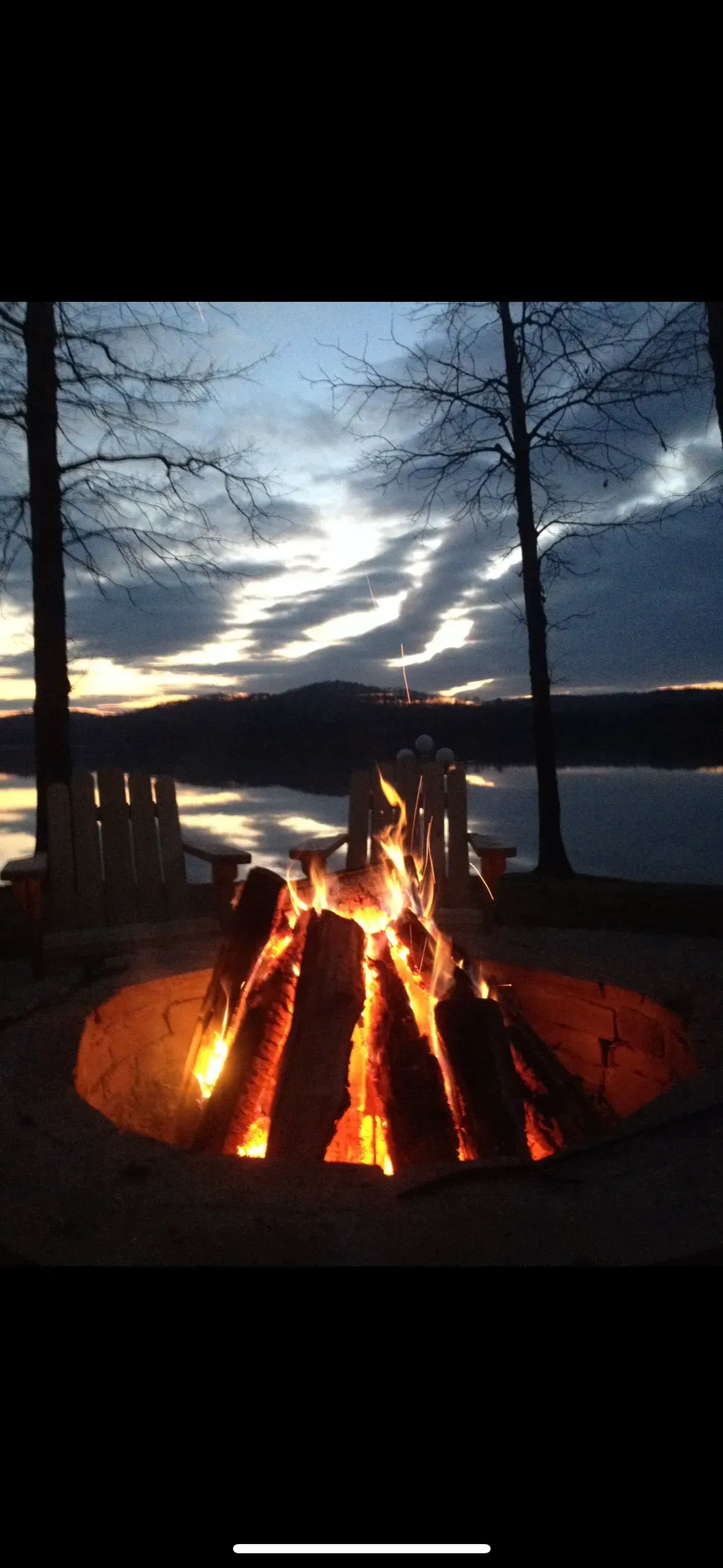 A campfire burns brightly in a fire pit, with a lake and trees in the background at dusk.