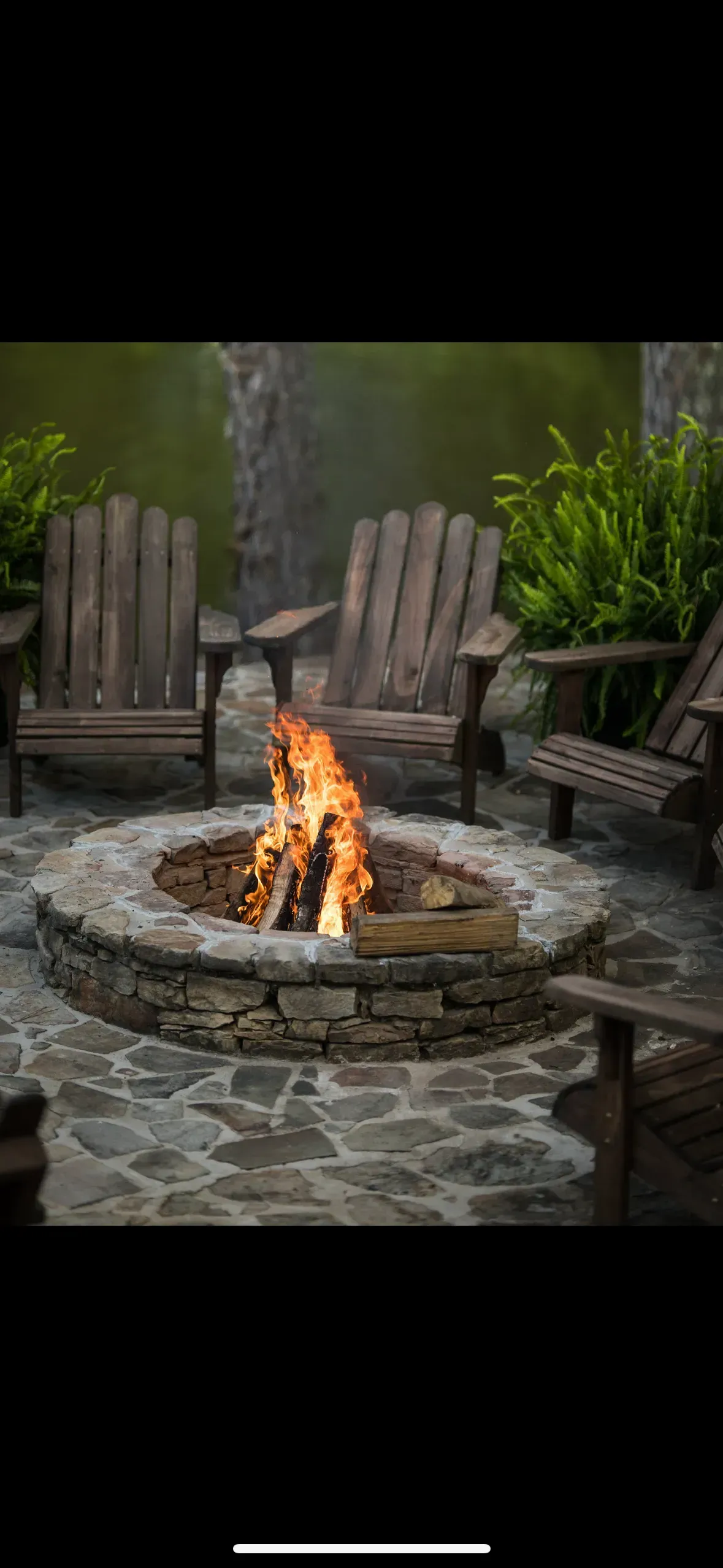 A fire pit surrounded by wooden chairs on a stone patio with greenery in the background.