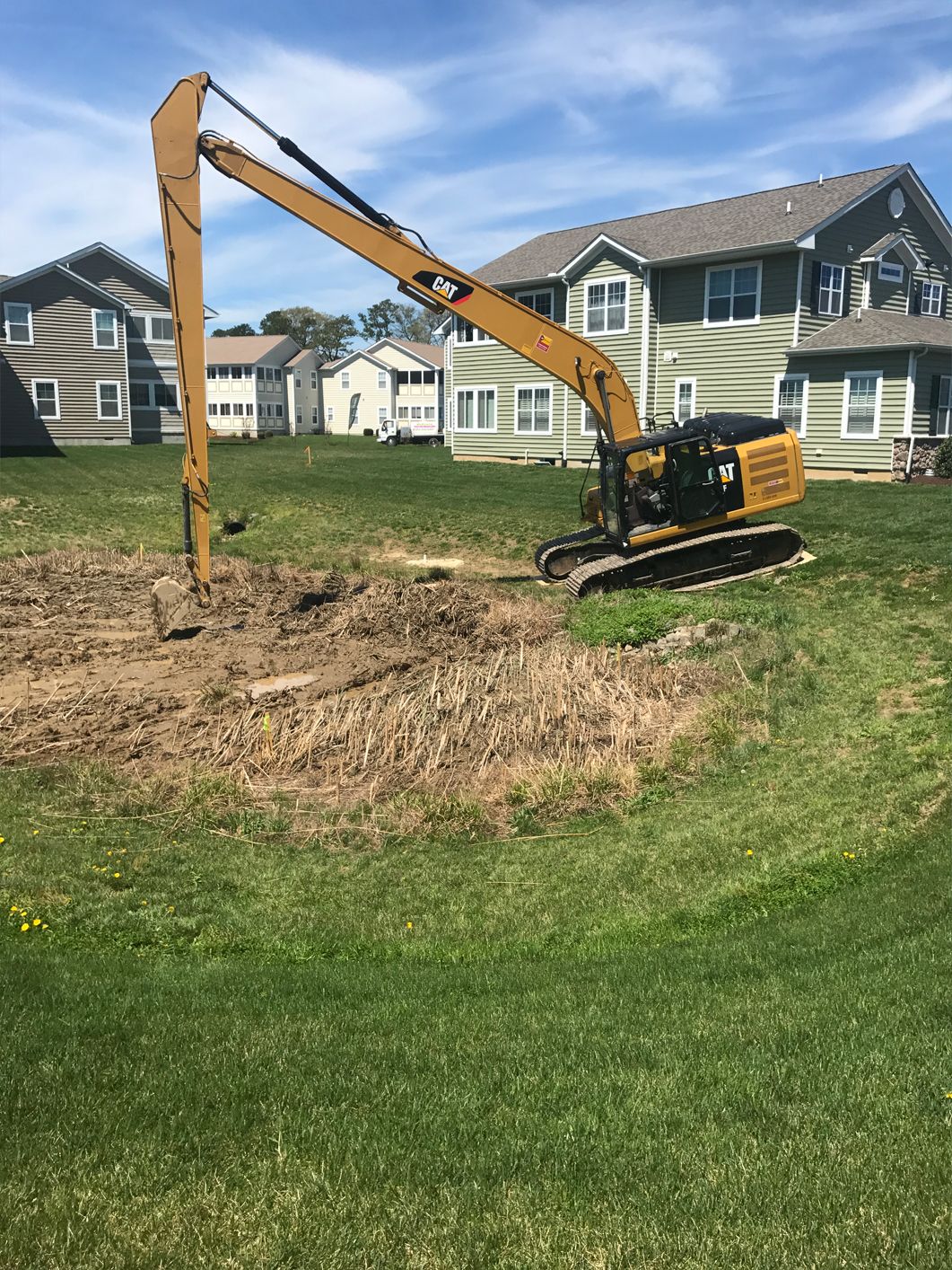A large yellow excavator is digging a hole in a grassy field in front of a house.