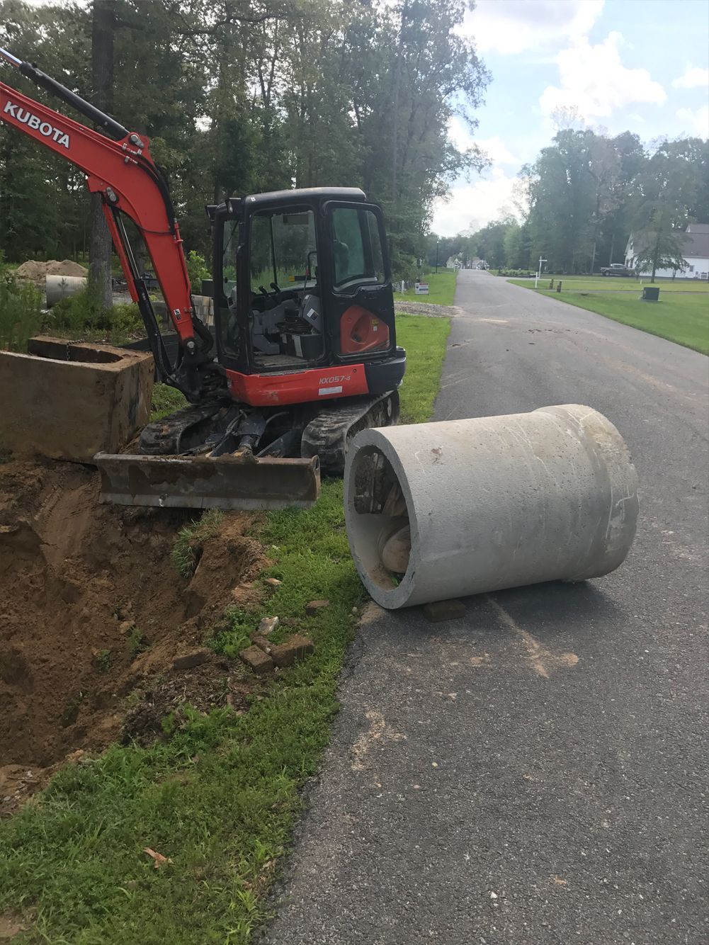 A large concrete pipe is laying on the side of the road next to an excavator.