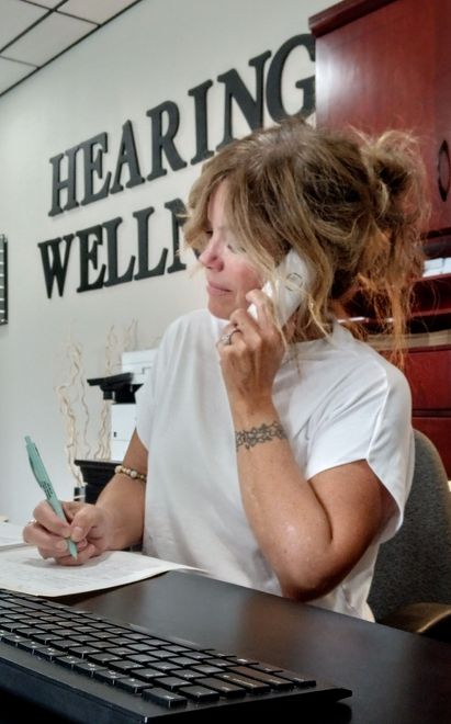 A woman is sitting at a desk in front of a computer in a hearing wellness office.