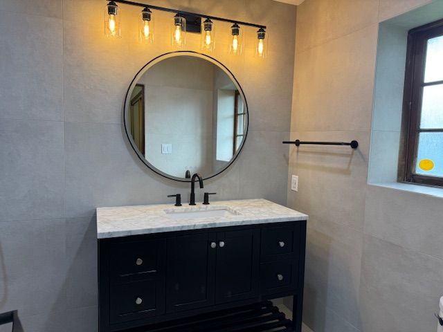 Bathroom with black vanity, round mirror, and overhead light fixture, on gray tiled walls.