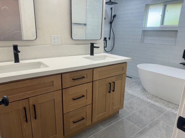 Modern bathroom with wooden vanity, two sinks, black fixtures, and a freestanding tub.