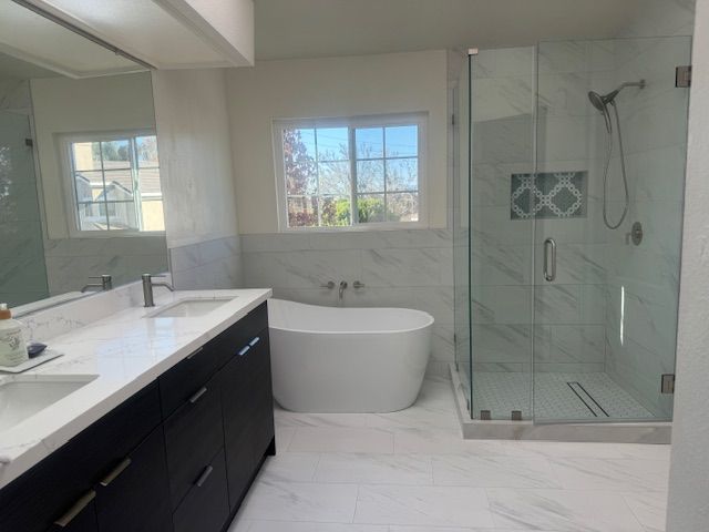 Bathroom with white marble tile, black vanity, free-standing tub, glass shower, and window.