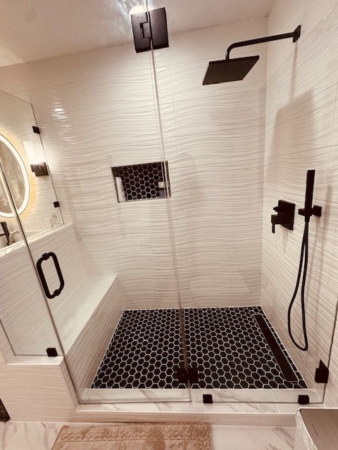 Modern bathroom shower with white textured tiles, black fixtures, glass doors, and dark hexagonal floor tiles.