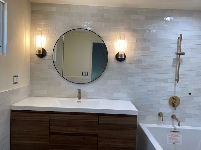 Bathroom with a wood vanity, round mirror, and wall sconces against a tile wall.