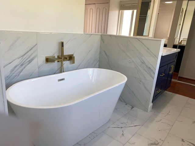 White freestanding bathtub with gold faucet against marble tile and blue vanity.