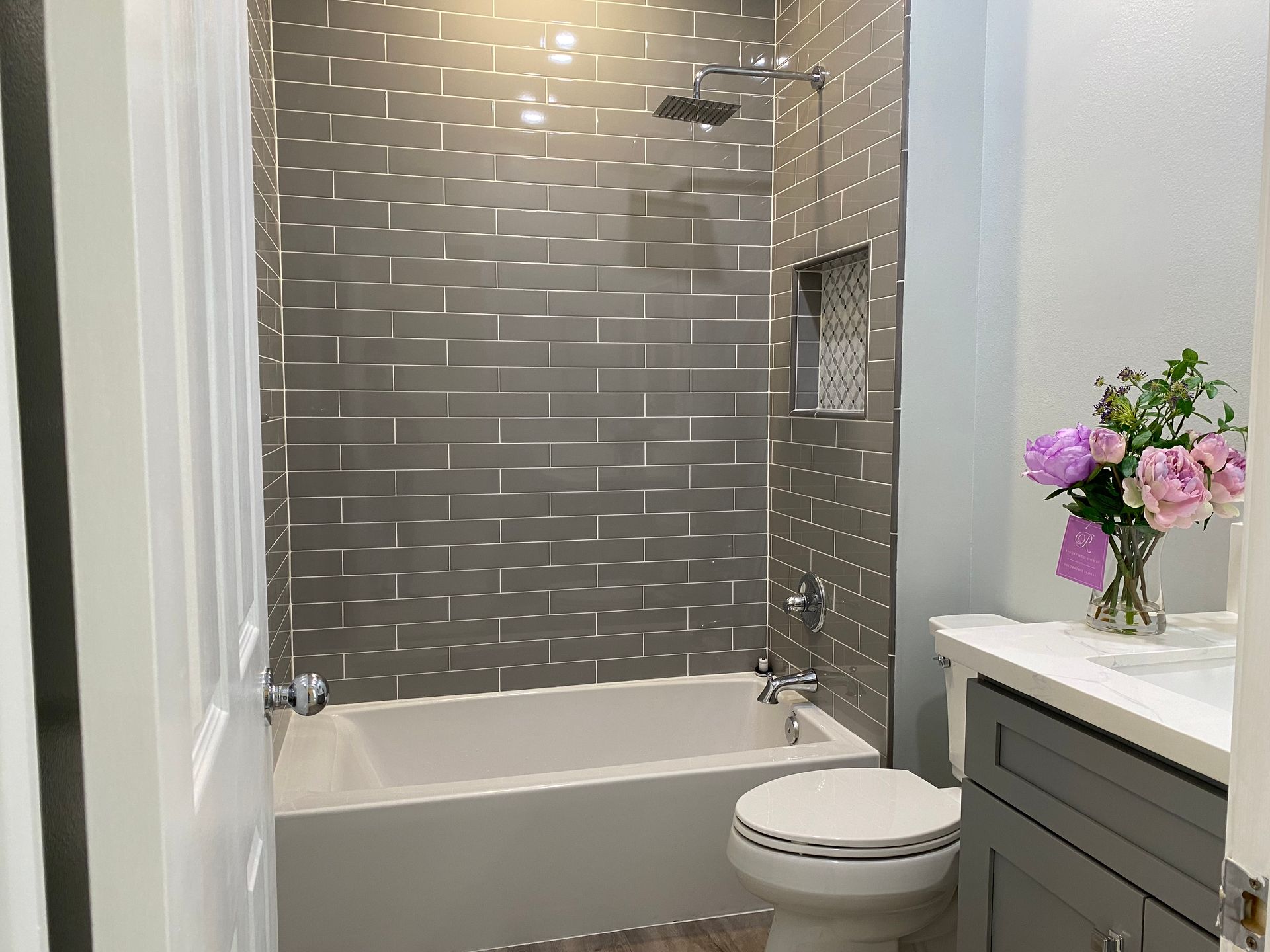 Bathroom with gray tiled shower, white tub and toilet, gray vanity, and a vase of flowers.
