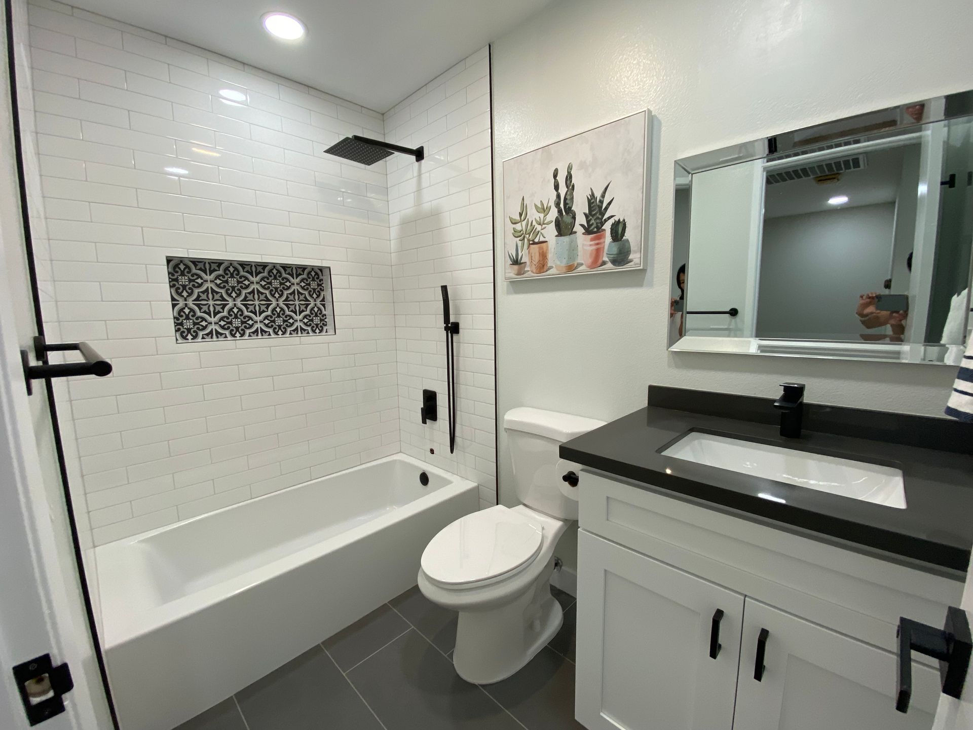 Modern bathroom: white subway tile shower, black fixtures, gray floor, white vanity with dark countertop, cactus art.