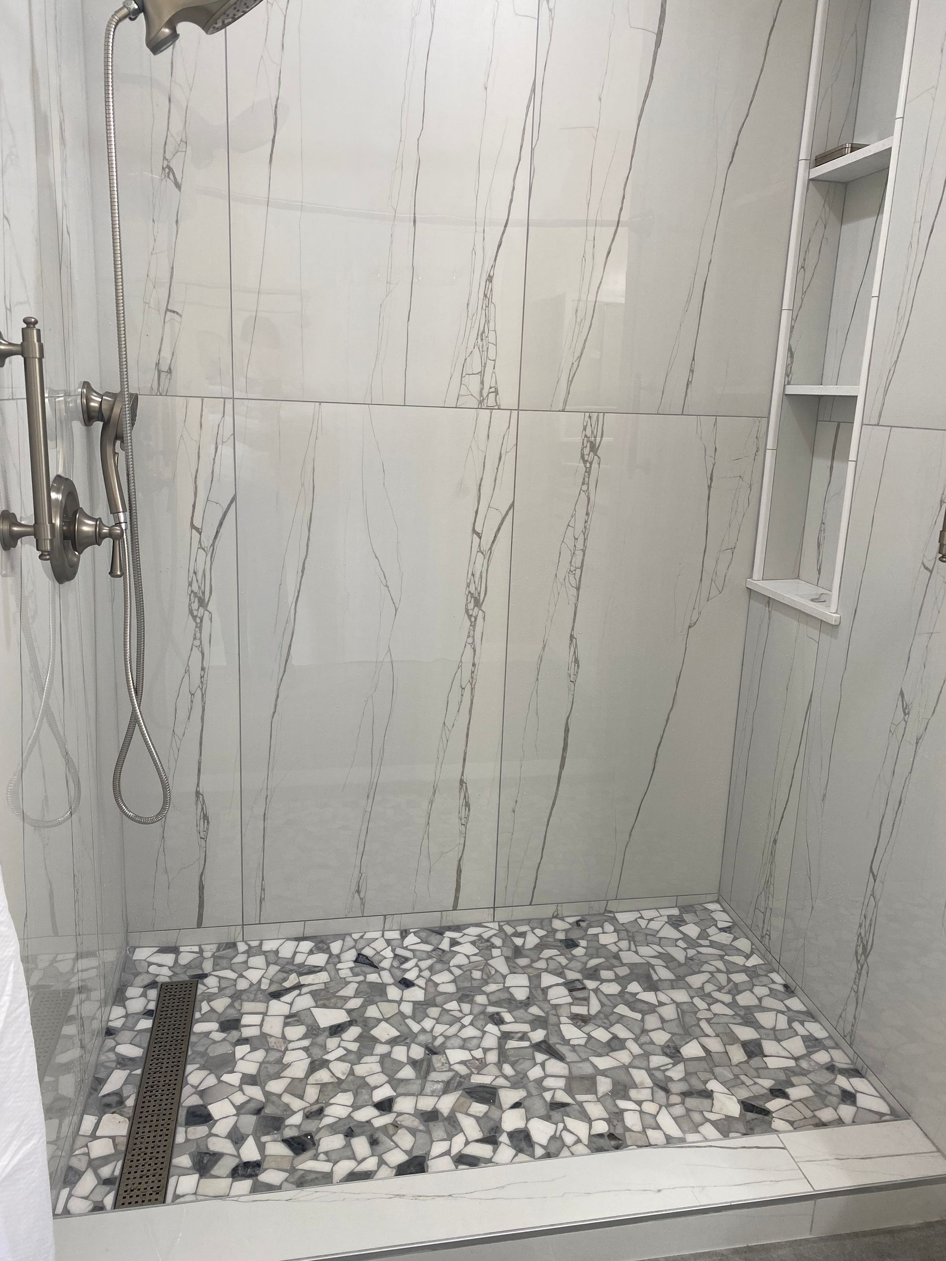 Shower stall with white and grey marble-look tiles, a mosaic floor, and built-in shelving.