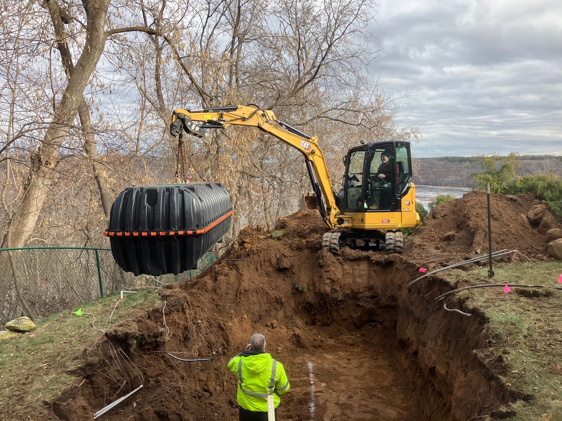 Yellow excavator lowers a septic tank into a trench, worker in neon vest oversees.