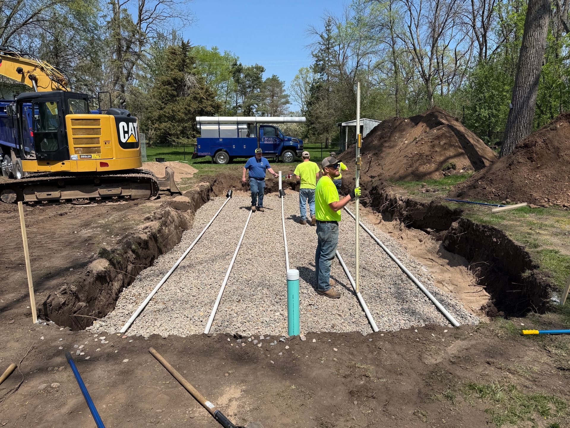 Construction site with workers installing pipes in a gravel-filled trench. A blue truck and excavator are visible.
