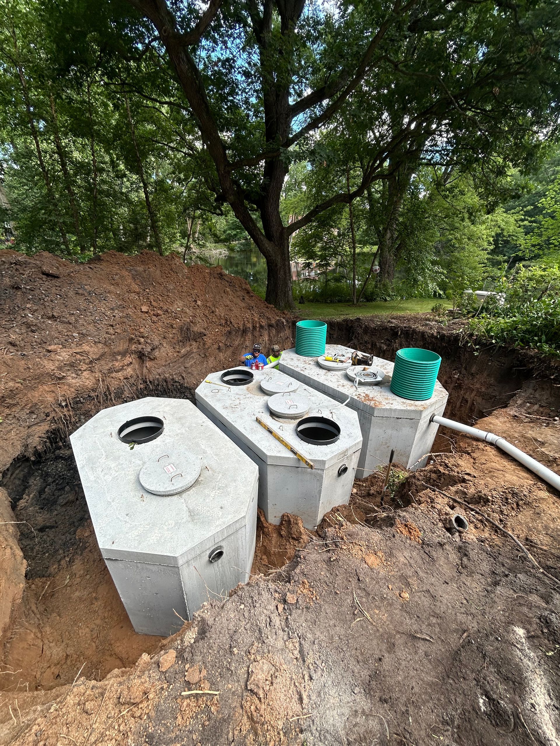 Three concrete septic tanks in a dirt excavation, under a tree, with green caps and a pipe.