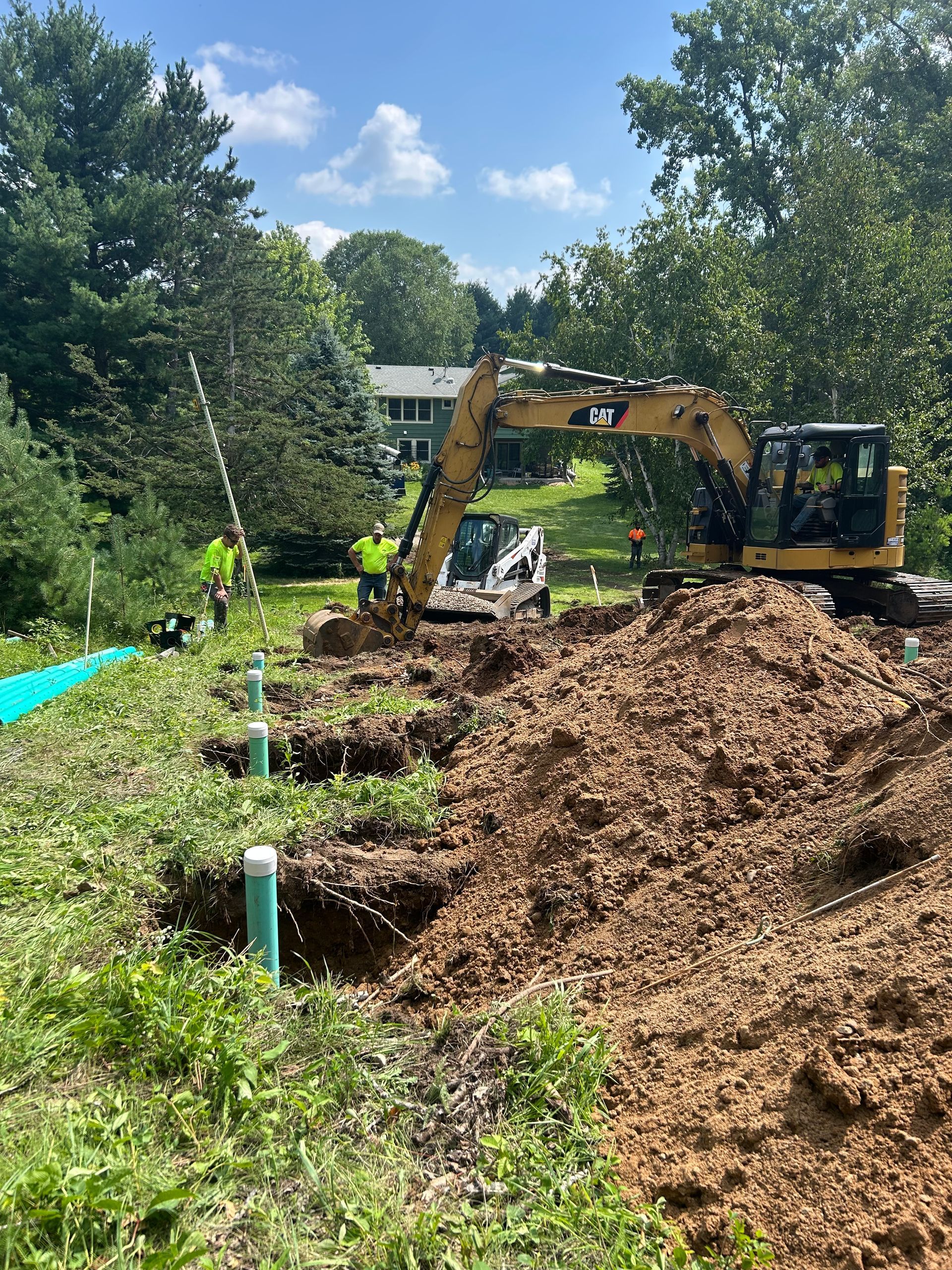 Construction site with a yellow excavator digging, two workers, and a small bulldozer in a green, wooded area.