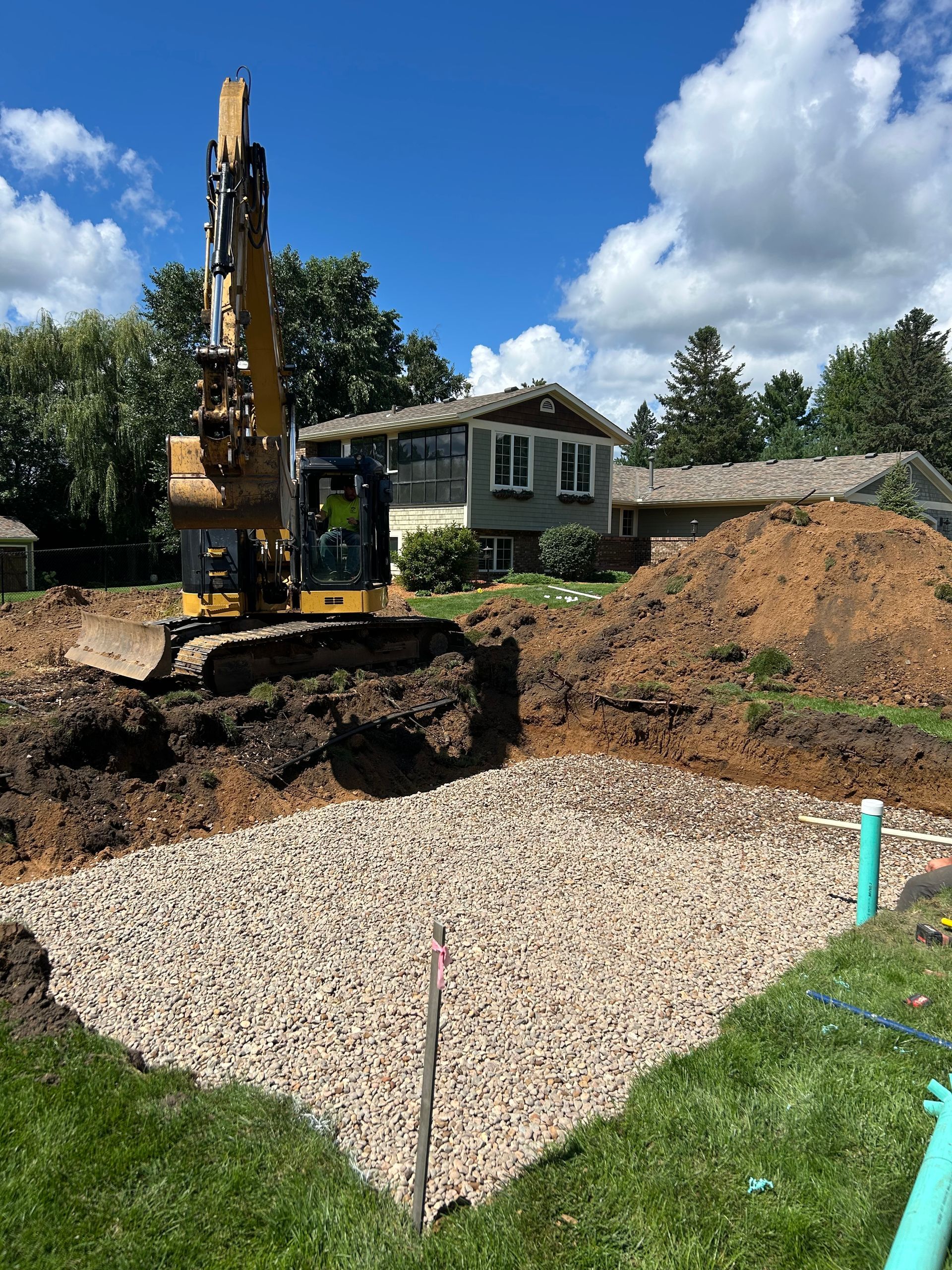 Excavator digging in yard, creating gravel-filled pit. Green grass, blue sky, houses in background.