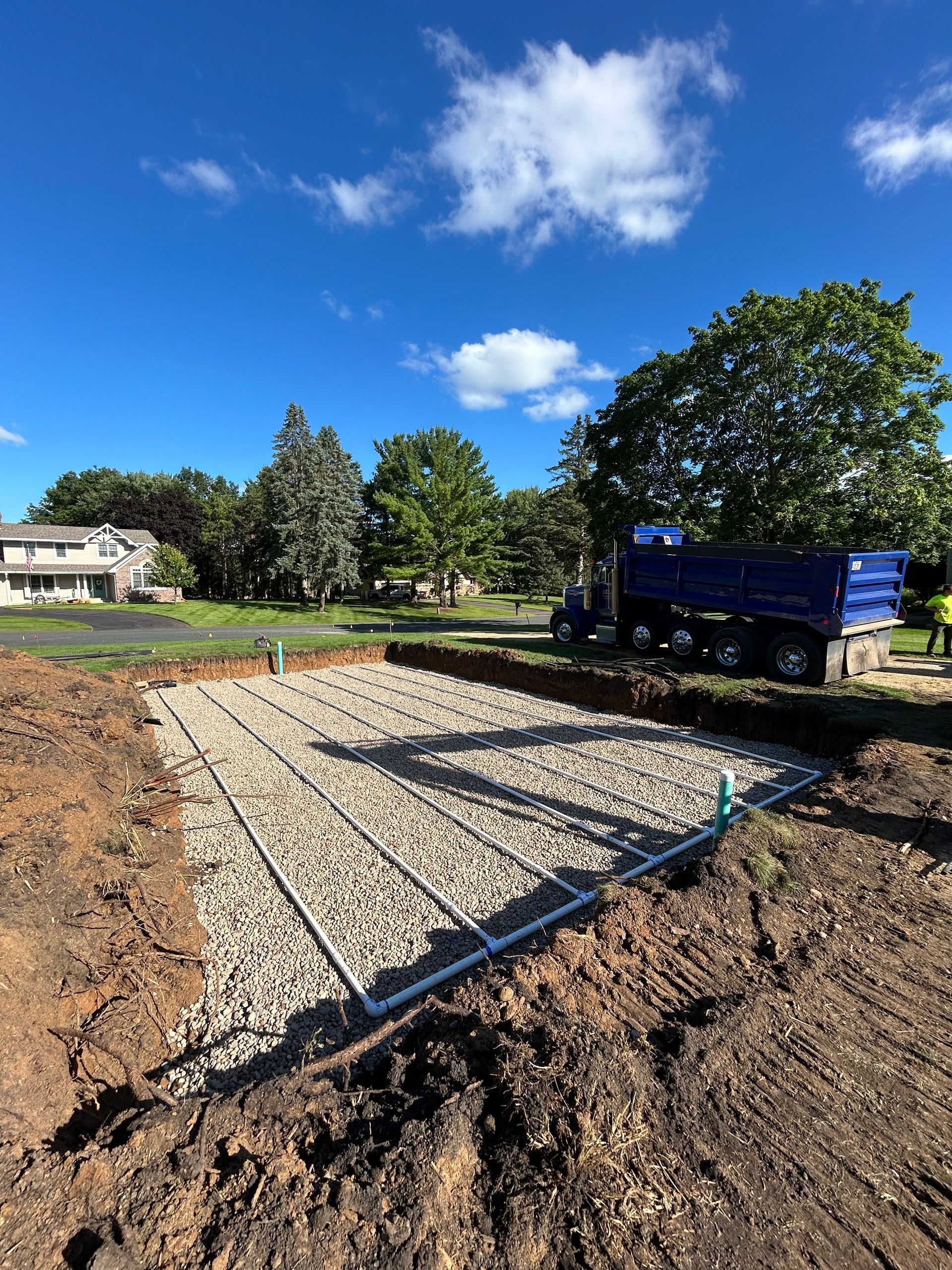 Excavated area with gravel and pipes, likely a septic system, near a dump truck under blue sky.