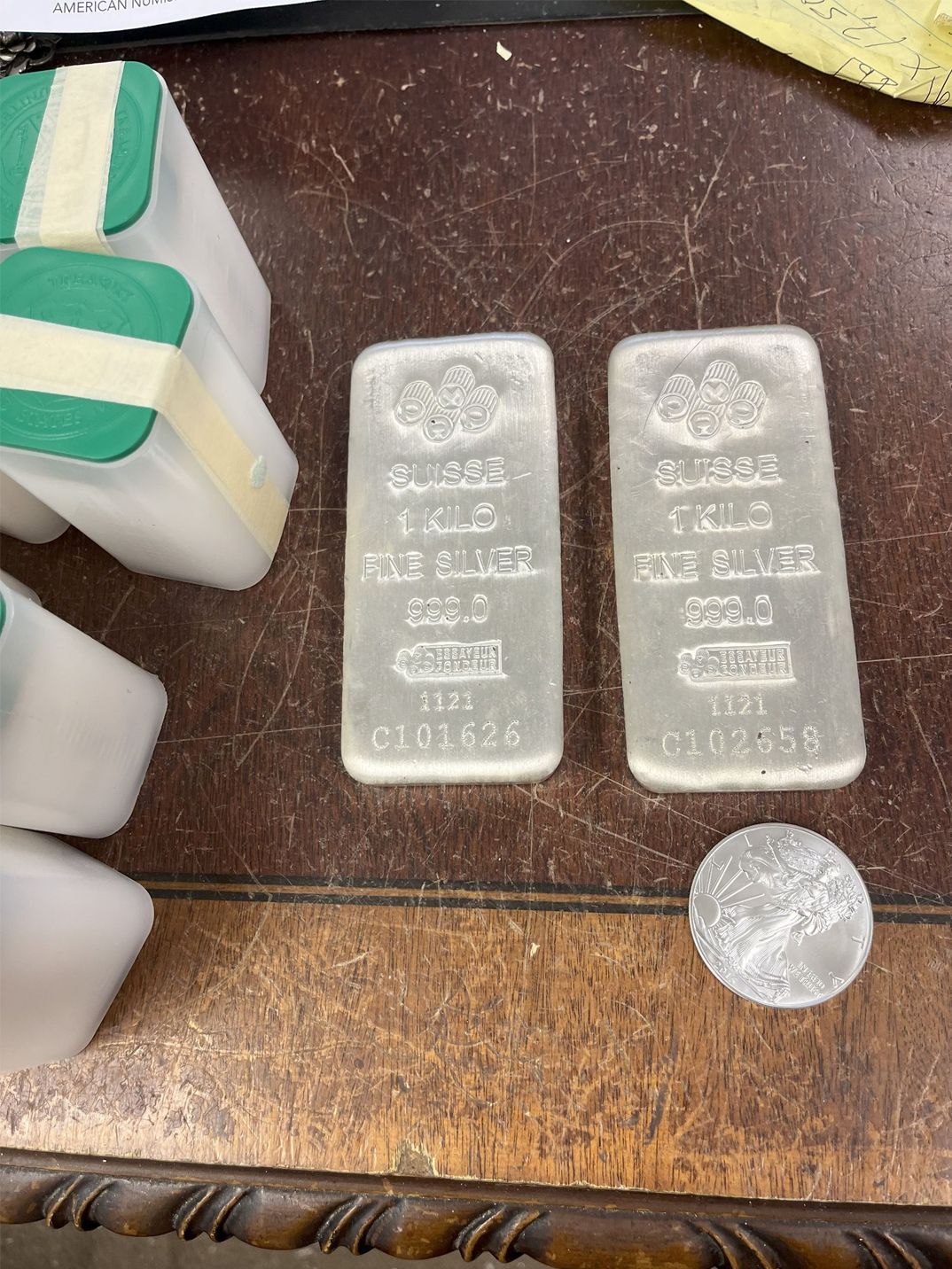 Two silver bars and a coin on a brown surface next to several tubes of coins.