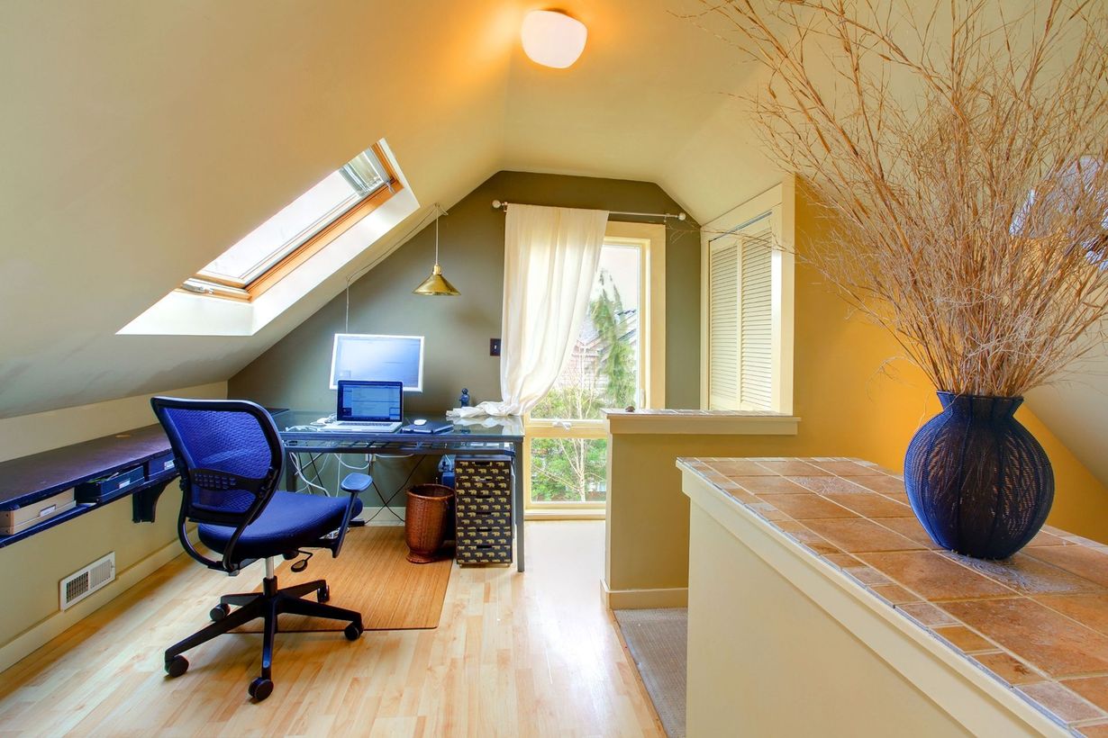 Bright home office with a blue chair, desk, skylight, and vase of dried branches by a window.