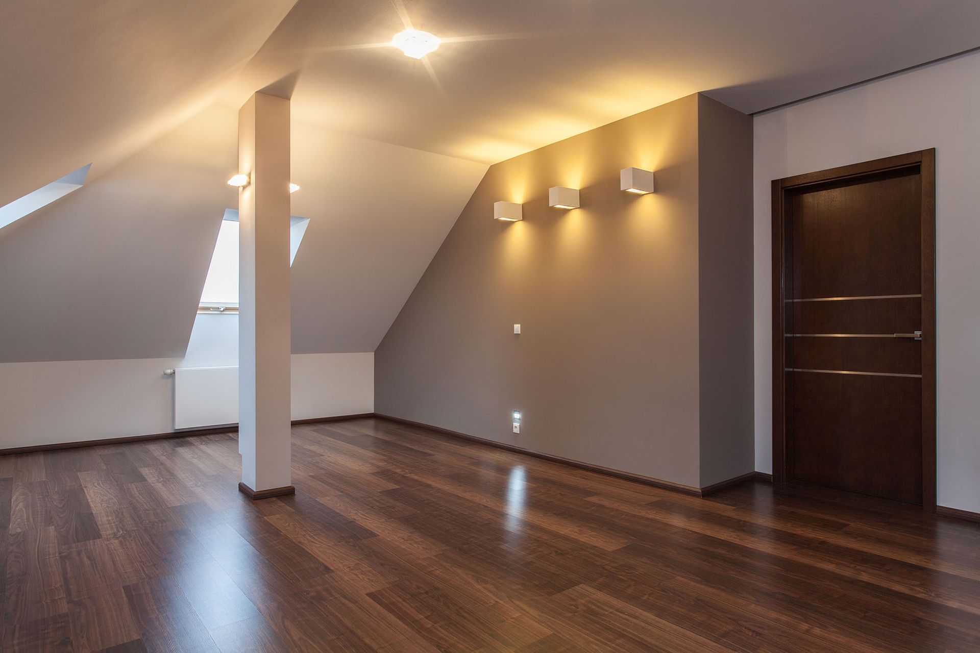 Empty attic room with hardwood floors, beige walls, and a dark brown door.