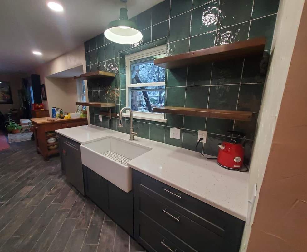A modern kitchen with dark cabinets, white countertops, and green tile backsplash. Floating wooden shelves above the sink.