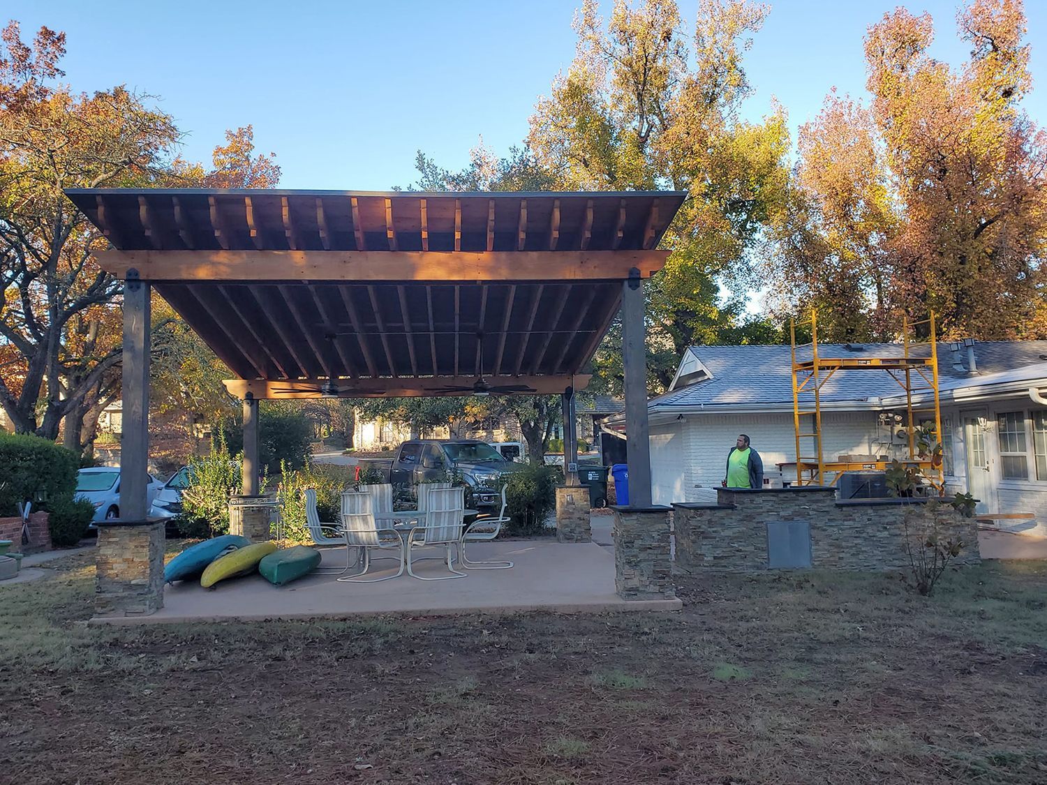 Pergola over a patio with seating, kayaks, and a small building. Autumn foliage surrounds the area.