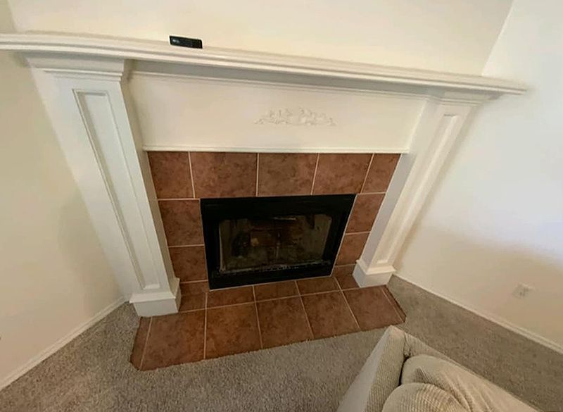 Fireplace with white mantel and brown tile surround, in a room with tan carpet.