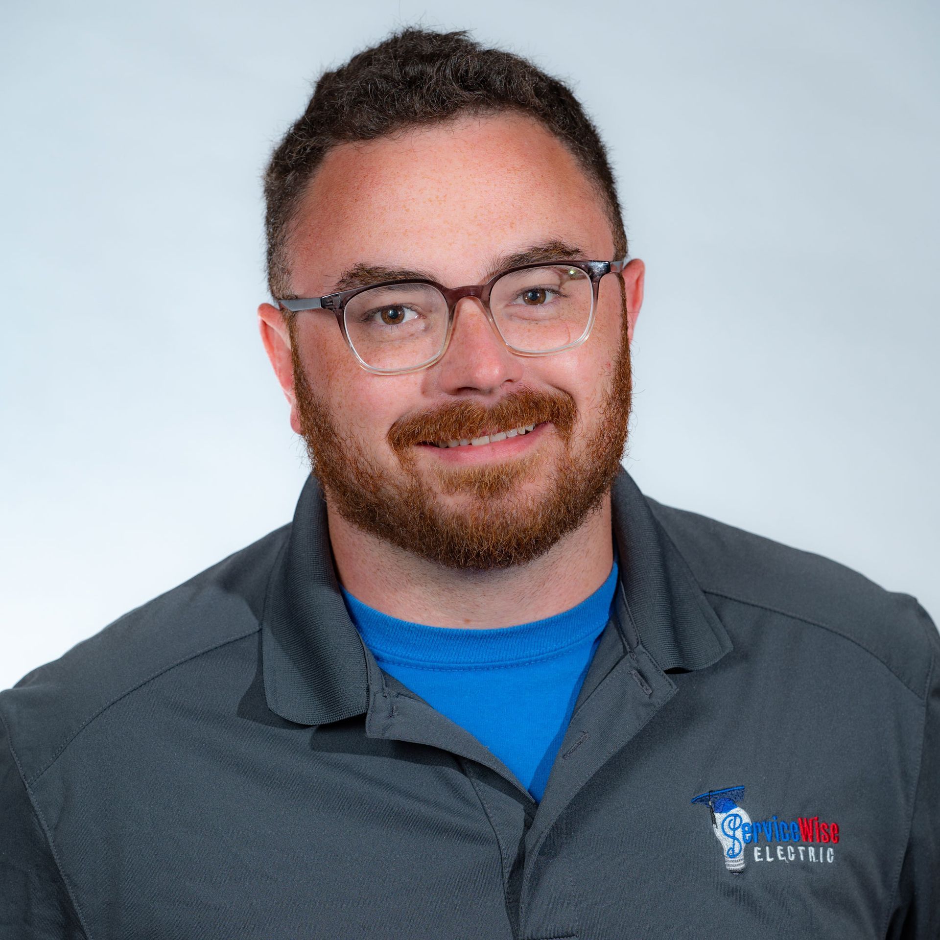 Man with glasses and beard smiles, wearing a gray polo over a blue shirt.