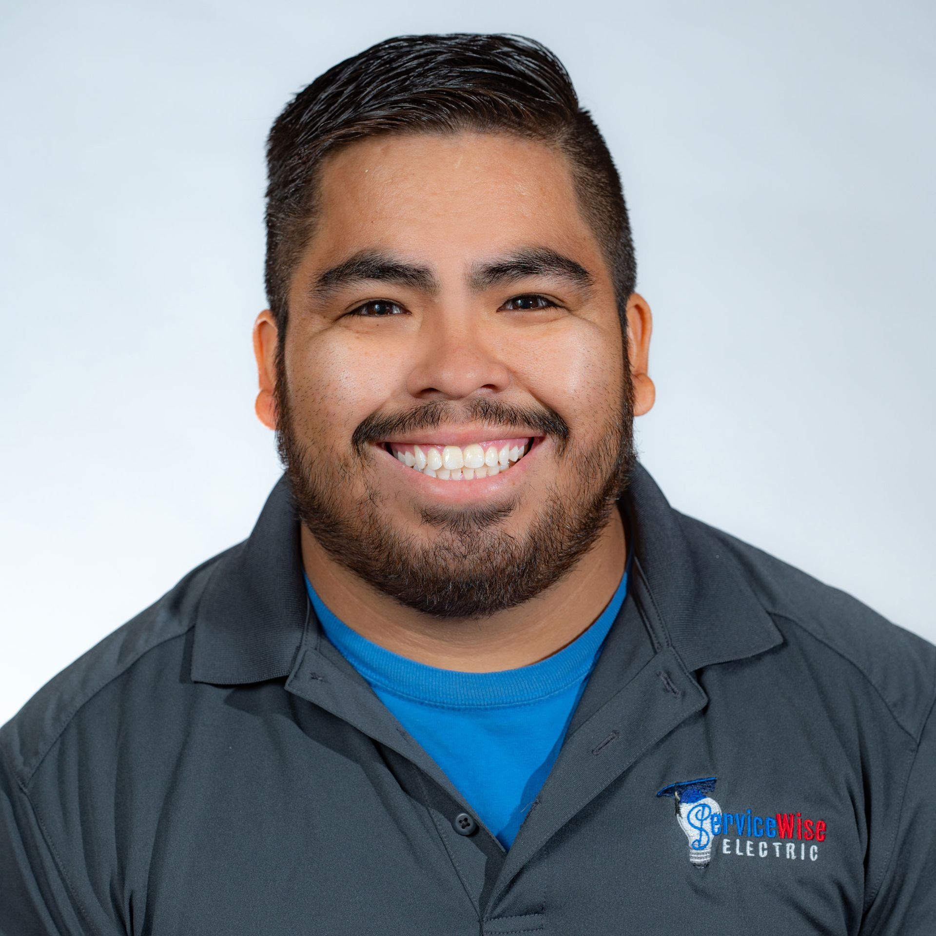 Smiling man in gray polo shirt, blue undershirt, logo on left chest, against white background.