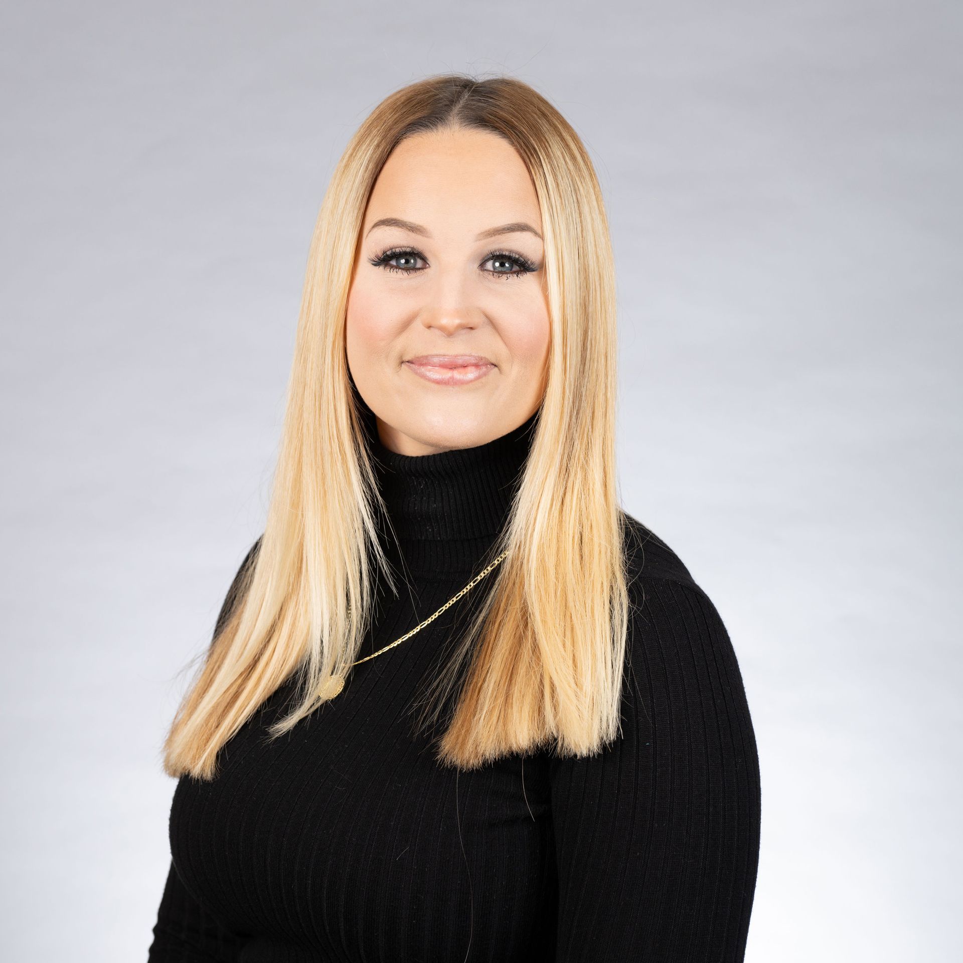 Woman with blonde hair wearing a black turtleneck and gold necklace, smiling, against a gray background.