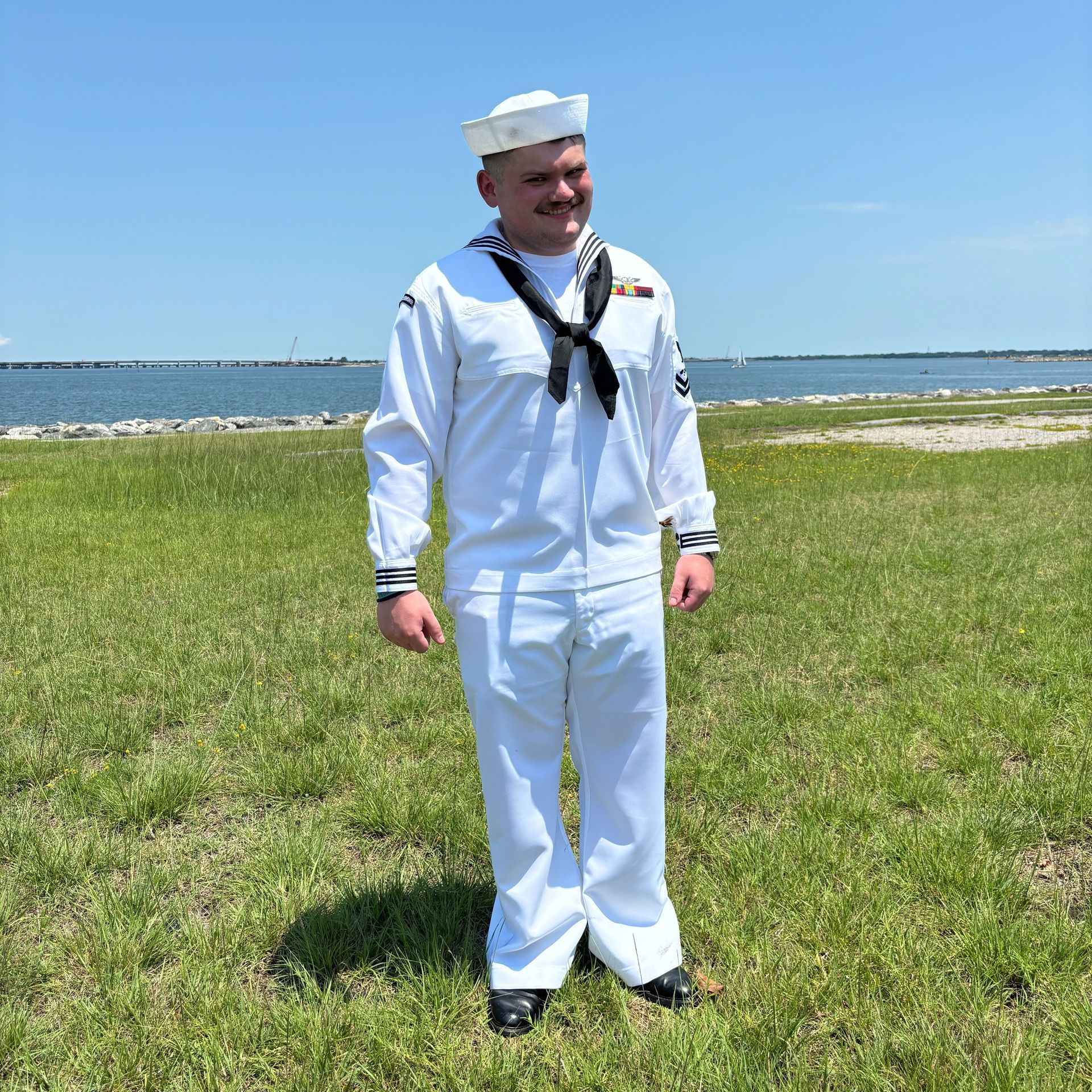 Sailor in white uniform stands on grassy field near water. Sunny, blue sky.
