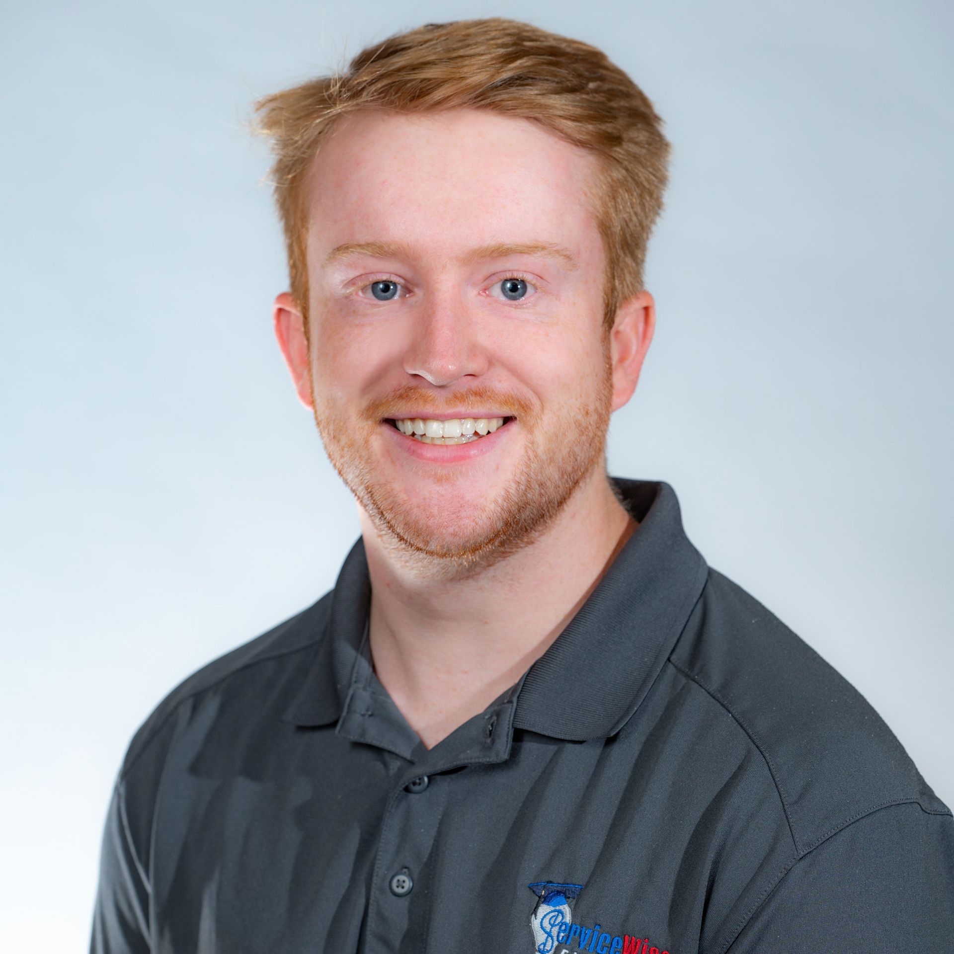 Man with red hair, smiling, wearing a gray polo shirt with a small emblem, set against a gradient background.